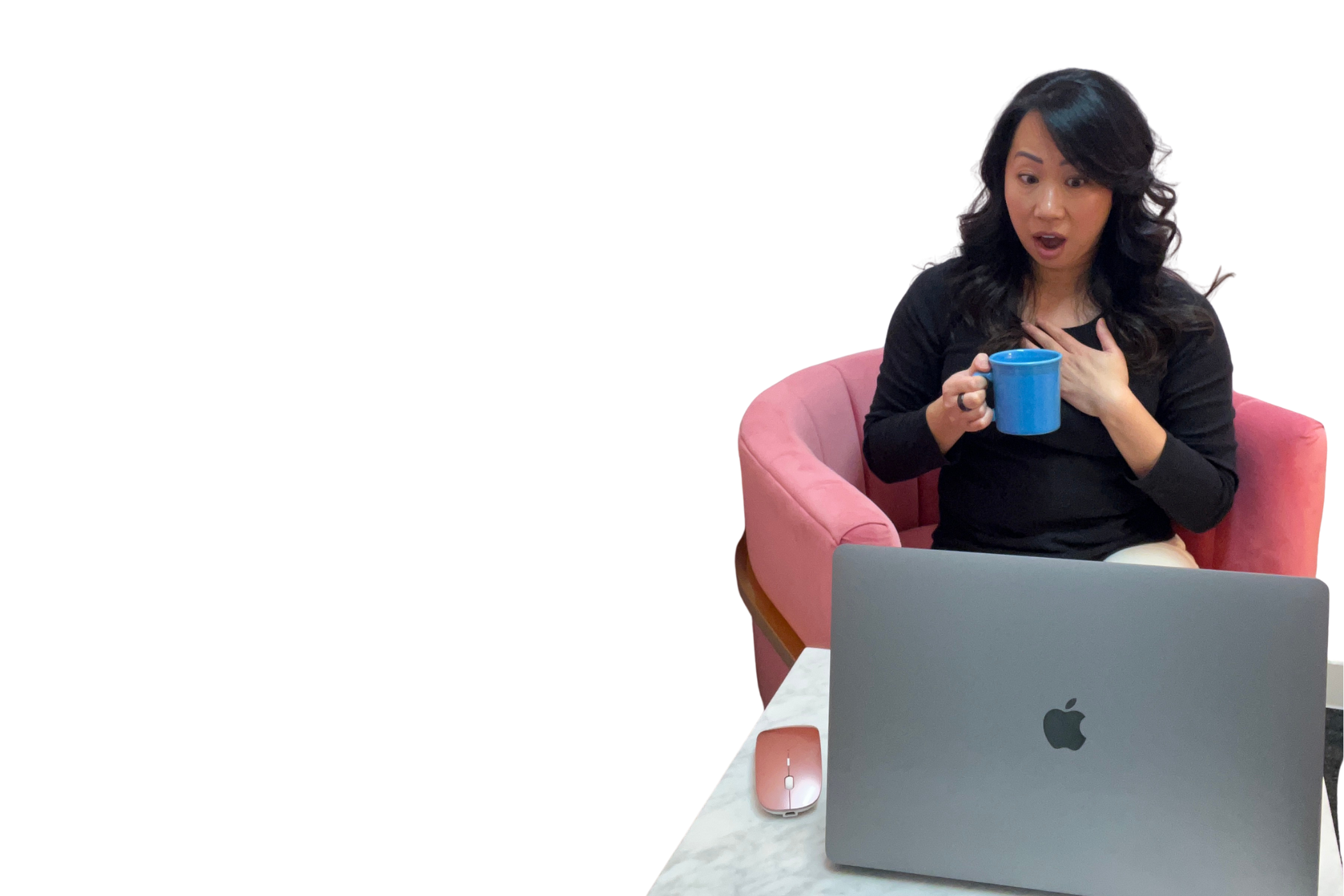 A woman sitting in a pink armchair holds a blue mug, with a silver MacBook on a white marble table in front of her.