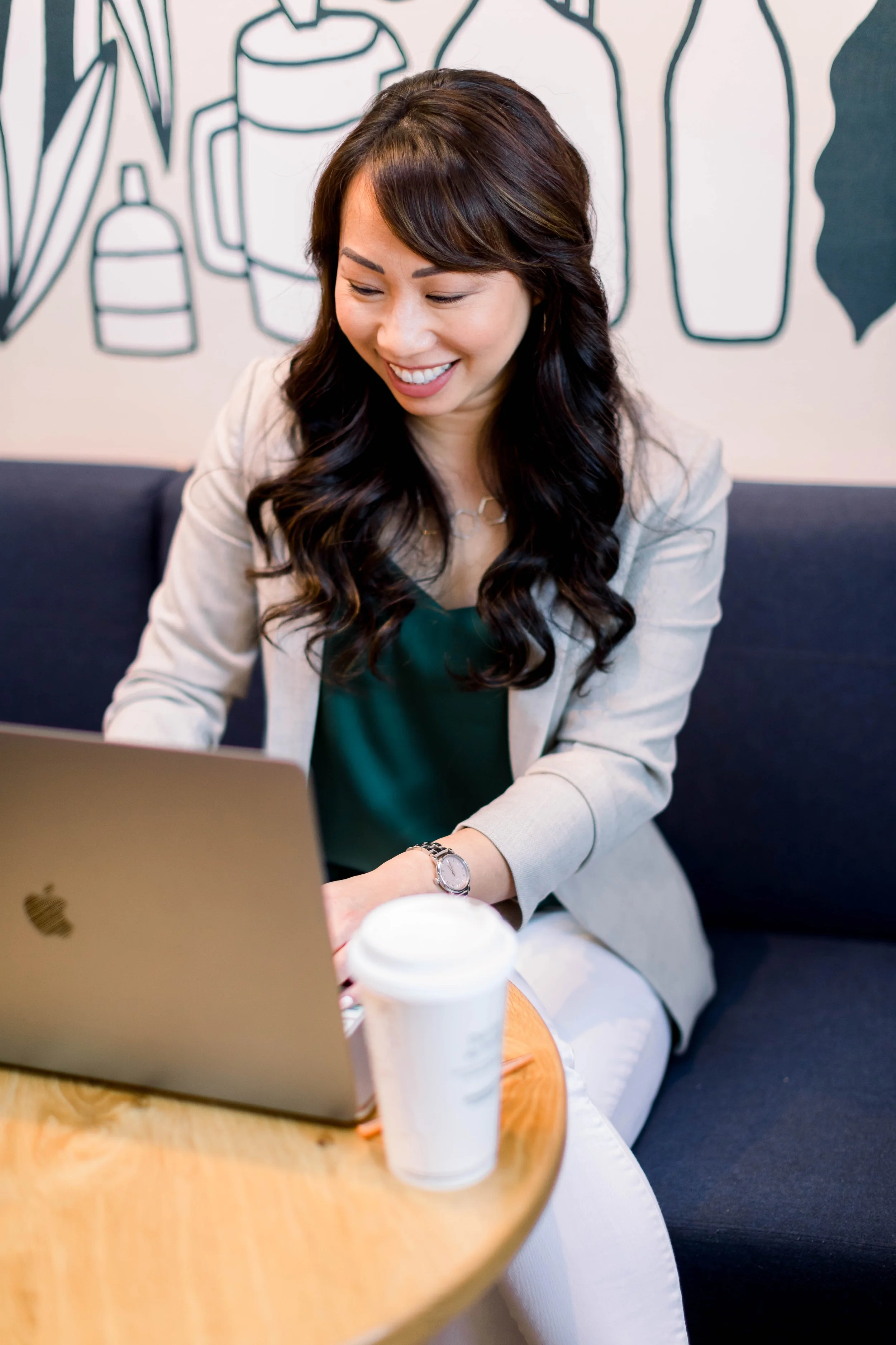 A woman with dark wavy hair and a beige blazer sitting at a cafe table with a MacBook laptop and a coffee cup, smiling, with a black and white line art mural on the wall behind her.