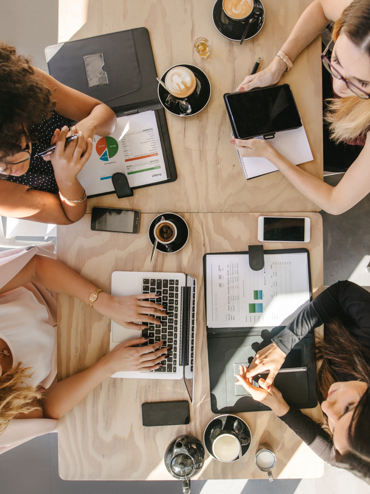 Top-down view of four women working at a wooden table with laptops, tablets, smartphones, coffee cups, and documents.