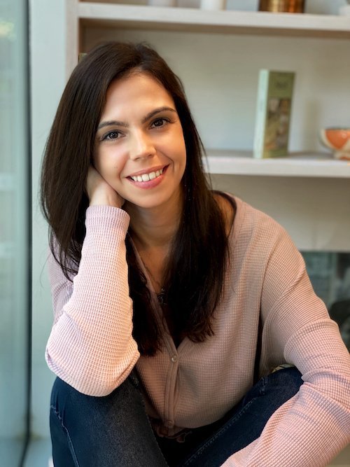 A woman with long dark hair, wearing a light pink sweater, is sitting indoors with a relaxed and happy expression.