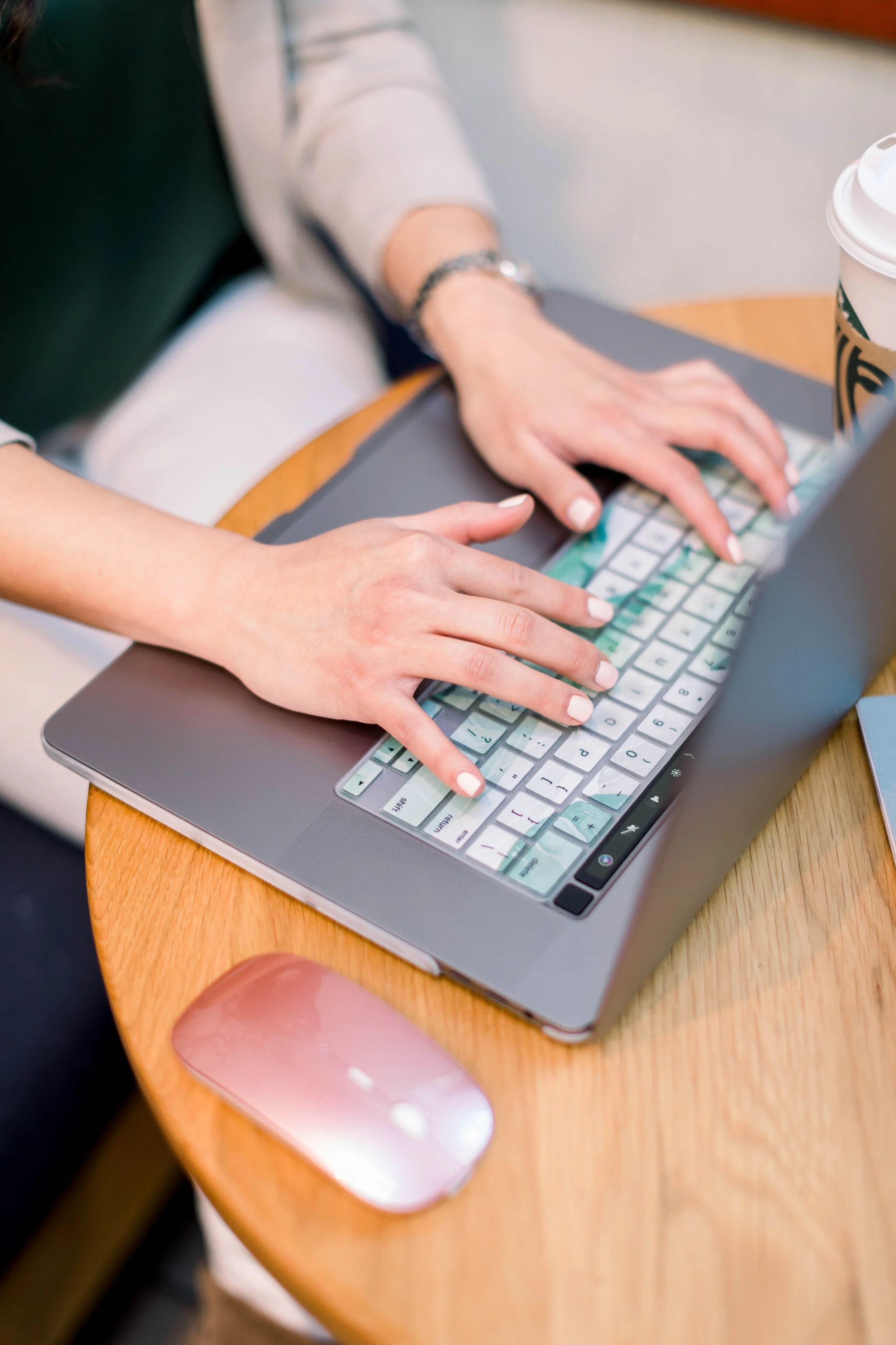 Person using a keyboard on a laptop at a wooden table with a pink computer mouse and a Starbucks cup.