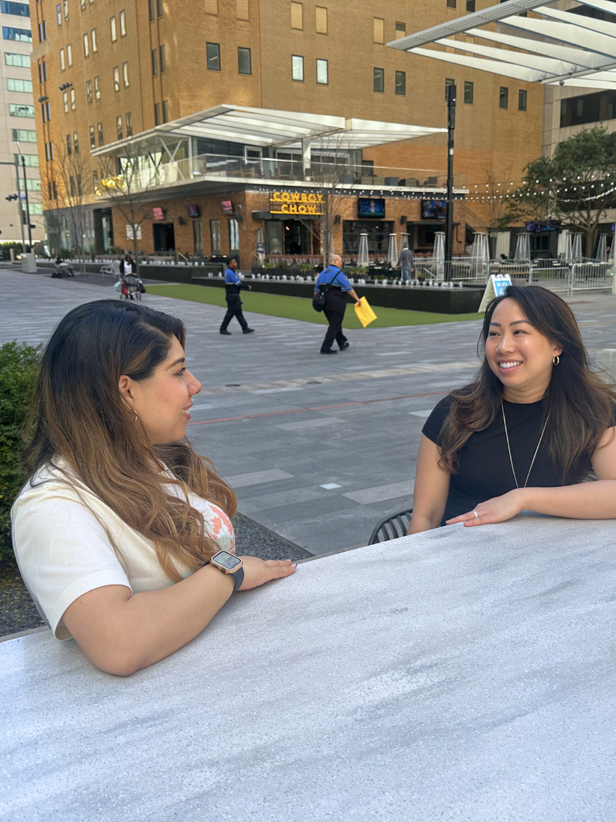 Two women are sitting at an outdoor table, smiling at each other, with a city street scene in the background that includes people walking, a building with a restaurant sign, and modern architecture.