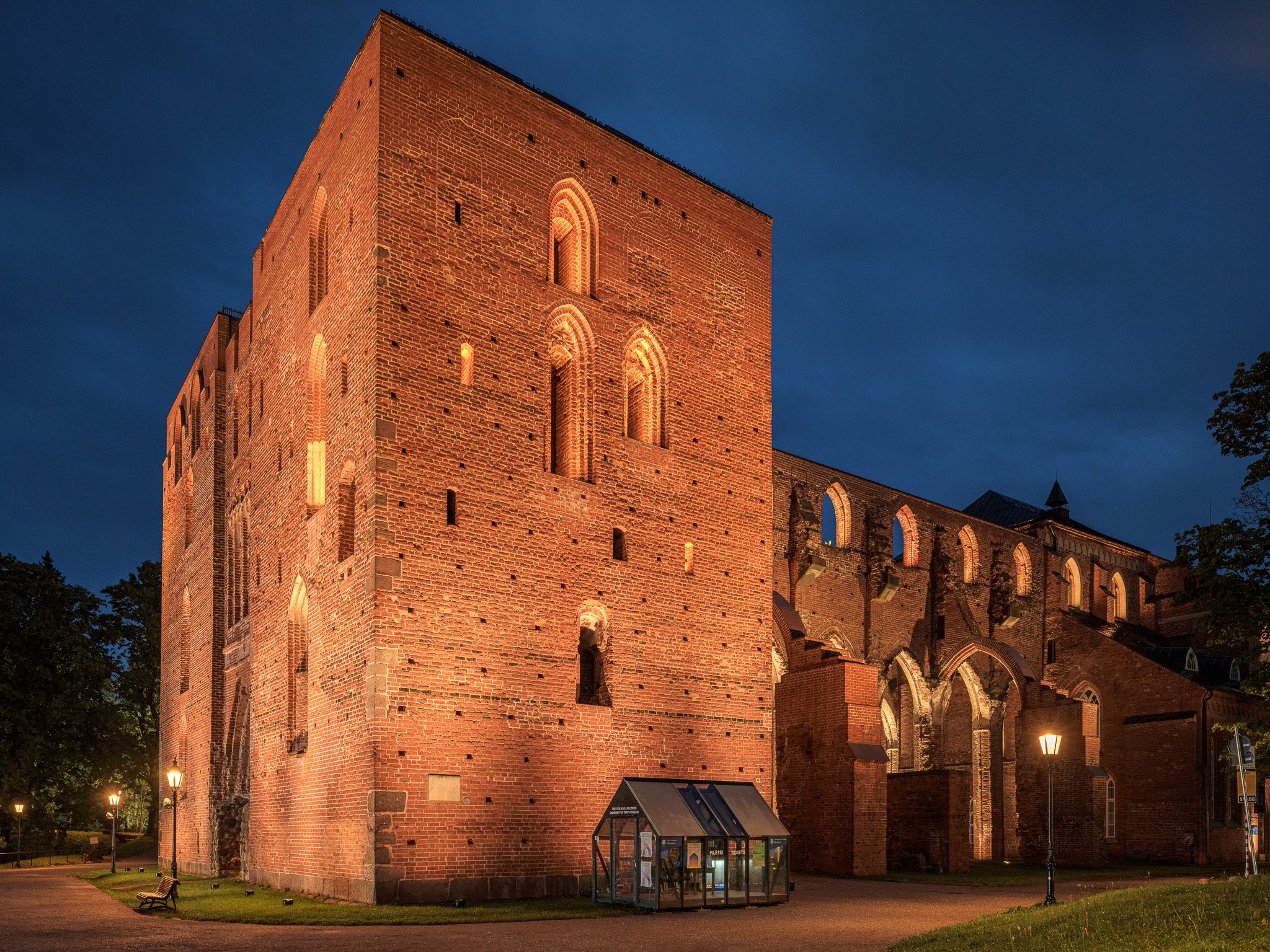 Vue nocturne d'un bâtiment en briques rouges avec des arcs et des fenêtres, éclairé par des lumières jaunes, entouré de lampadaires et de quelques arbres.