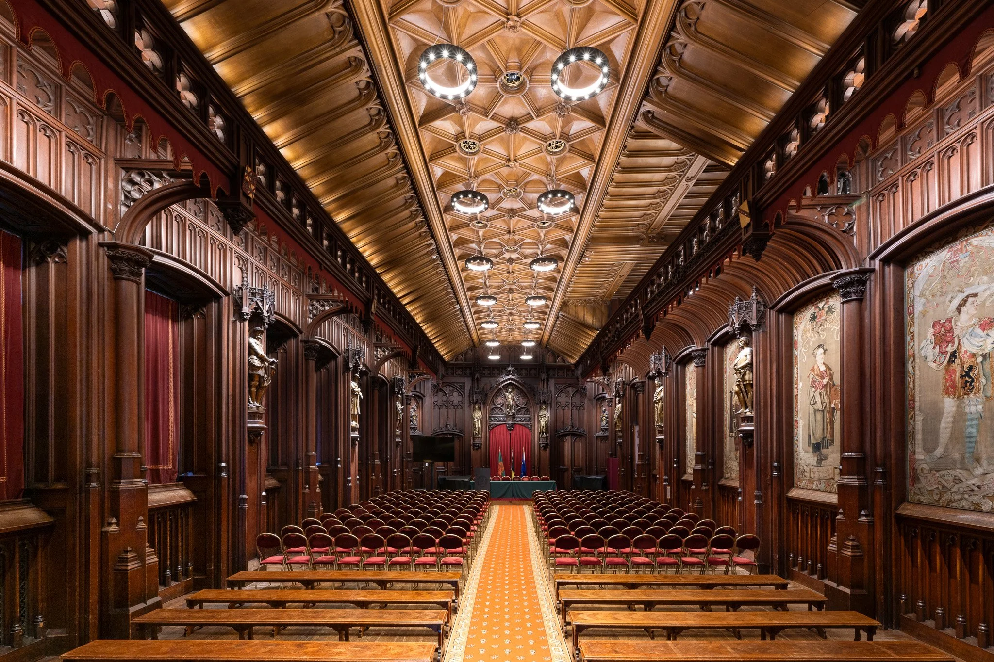 Grande salle ornée de boiseries sculptées, au plafond élaboré, avec des rangées de sièges rouges et un tapis central. Des tentures et statues décorent les murs.
