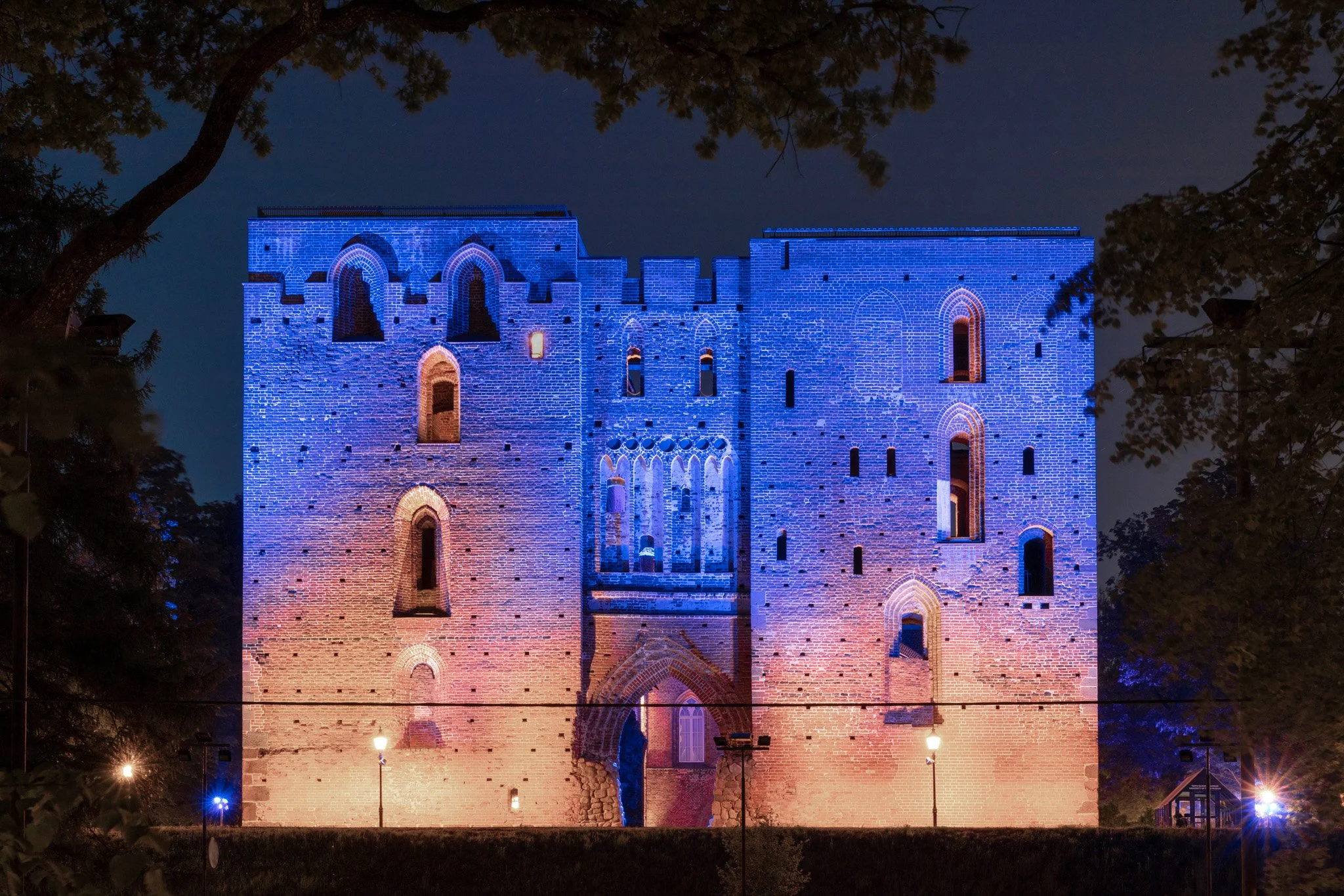 Château en briques illuminé en bleu et rouge la nuit, entouré d'arbres.