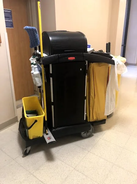 A cleaning cart with a mop, yellow trash bin, and cleaning supplies in a hospital or janitorial setting.