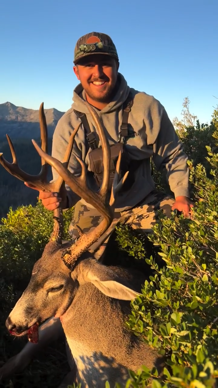 A smiling man in hunting gear holding the antlers of a freshly hunted deer with a mountainous landscape in the background.