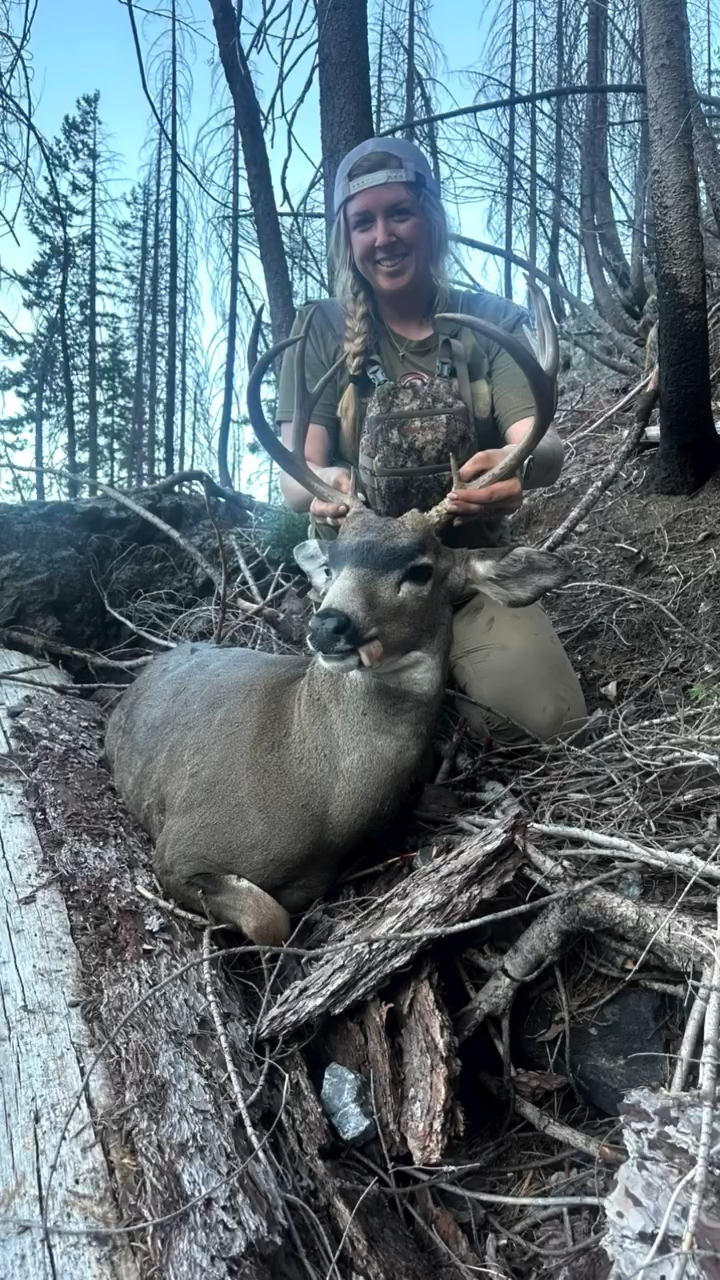 A woman with a braid, wearing a cap backwards, smiling, kneeling on the ground in a forest, holding the antlers of a deer with closed eyes and tongue sticking out, which is lying on the ground surrounded by branches and trees.