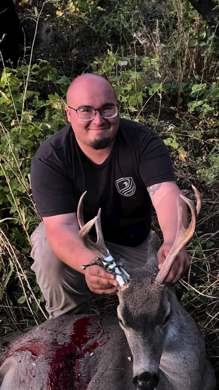 A man smiling and kneeling in a wooded area, holding a deceased deer with antlers, blood visible on the deer's side.