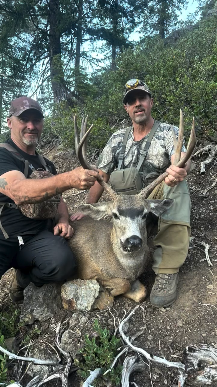 Two men posing with a large deer, holding its antlers in a forested area.