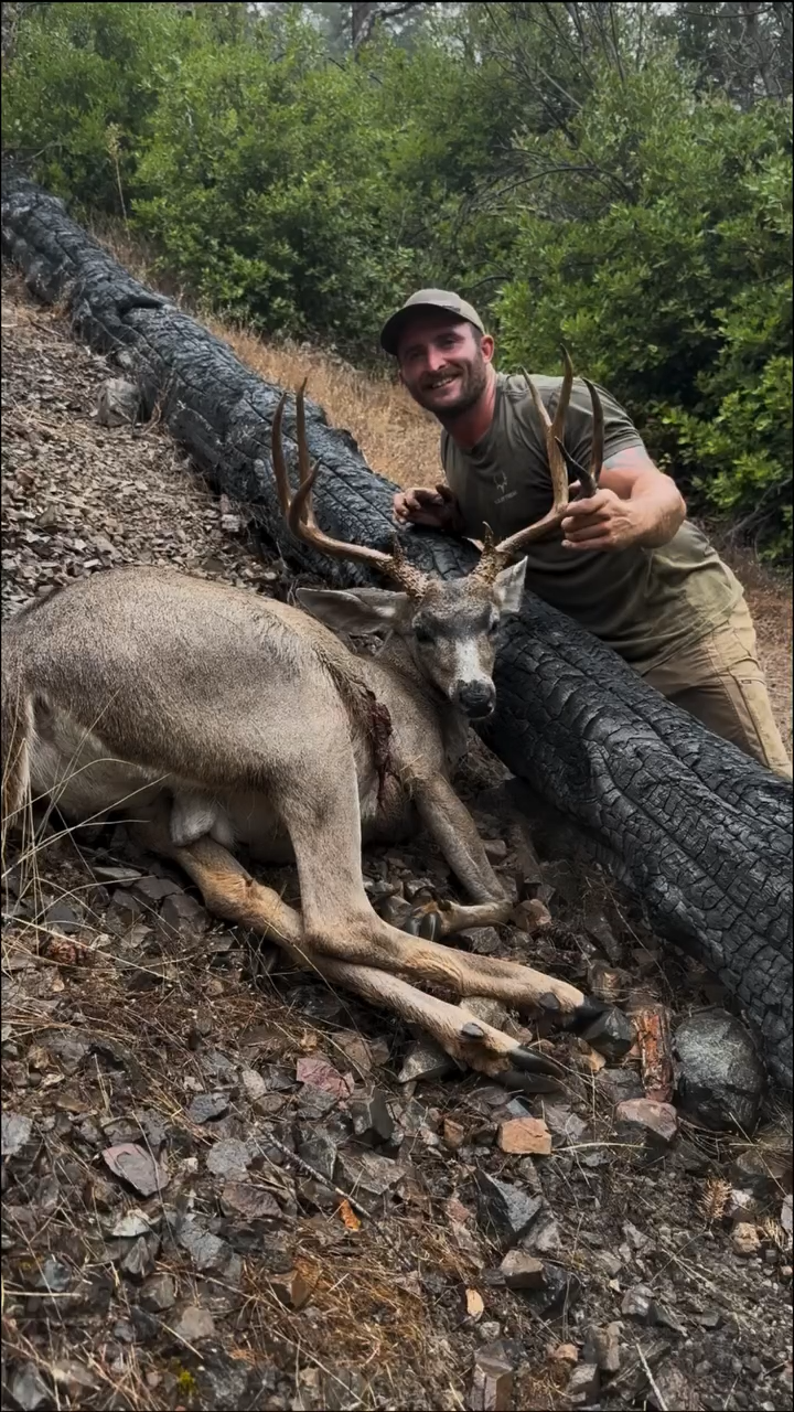 A man in outdoor gear leaning on a fallen tree beside a dead deer with large antlers.