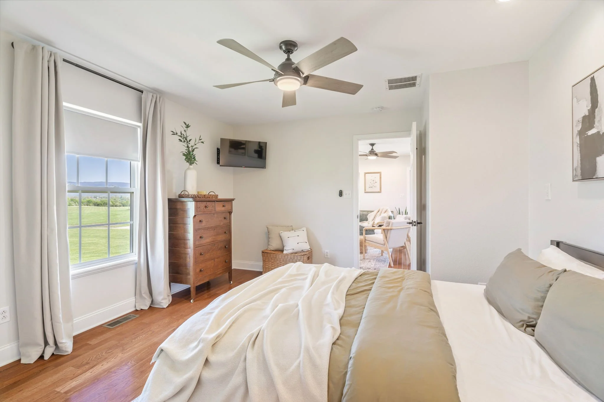 A bedroom with a bed, wooden dresser, wall-mounted TV, window with white curtains, and an open door leading to a sitting area with another ceiling fan and armchair.