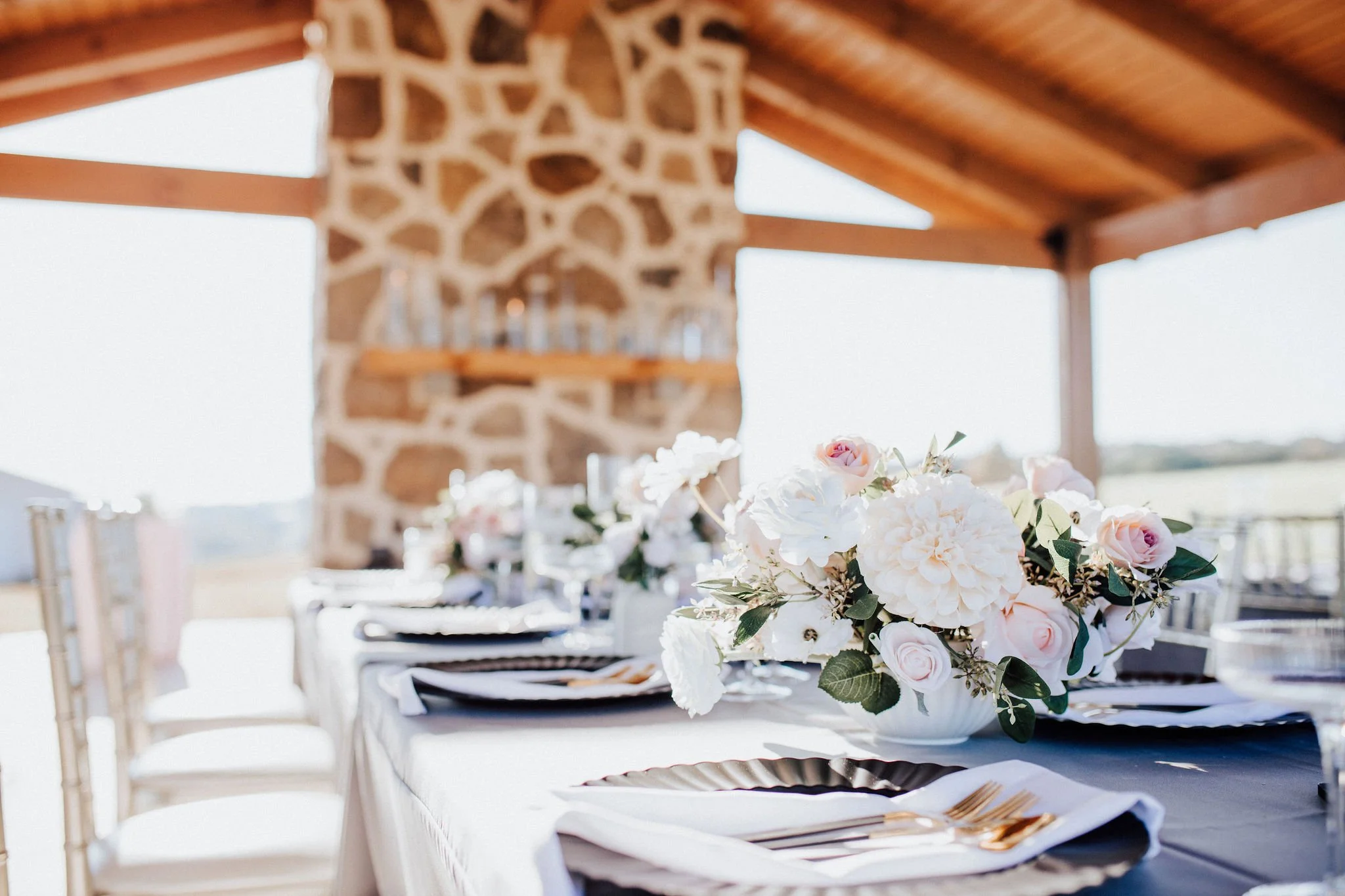 Elegant outdoor dining table with floral centerpiece, gold cutlery, black and white plates, under a wooden pavilion with stone fireplace, set for a special occasion.