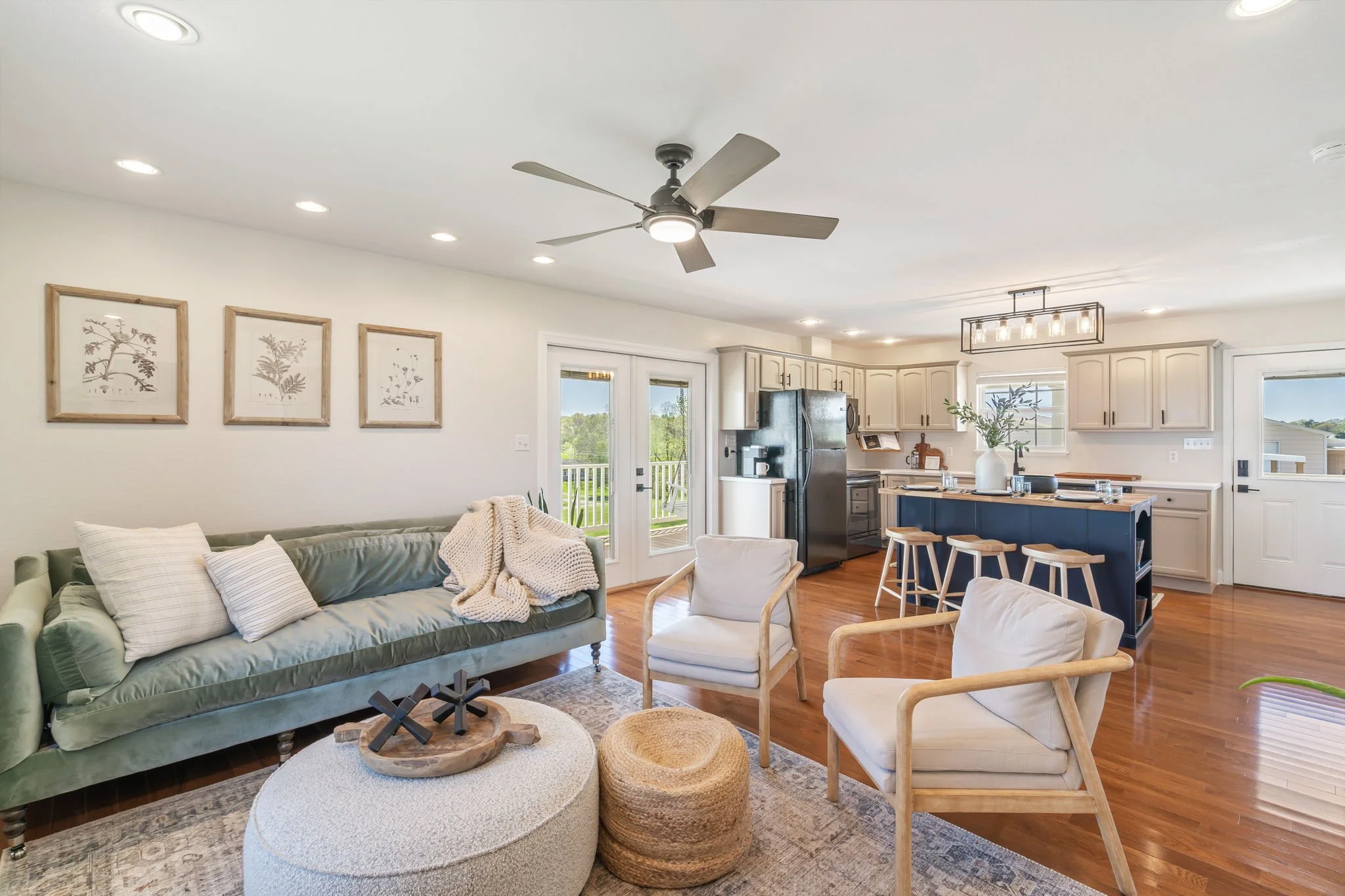 Open-concept living room and kitchen with a ceiling fan, beige sofa with pillows, white armchairs, round coffee table with decor, kitchen island with stools, black refrigerator, cabinet on white walls, hardwood floor, and sliding glass doors leading to a balcony.