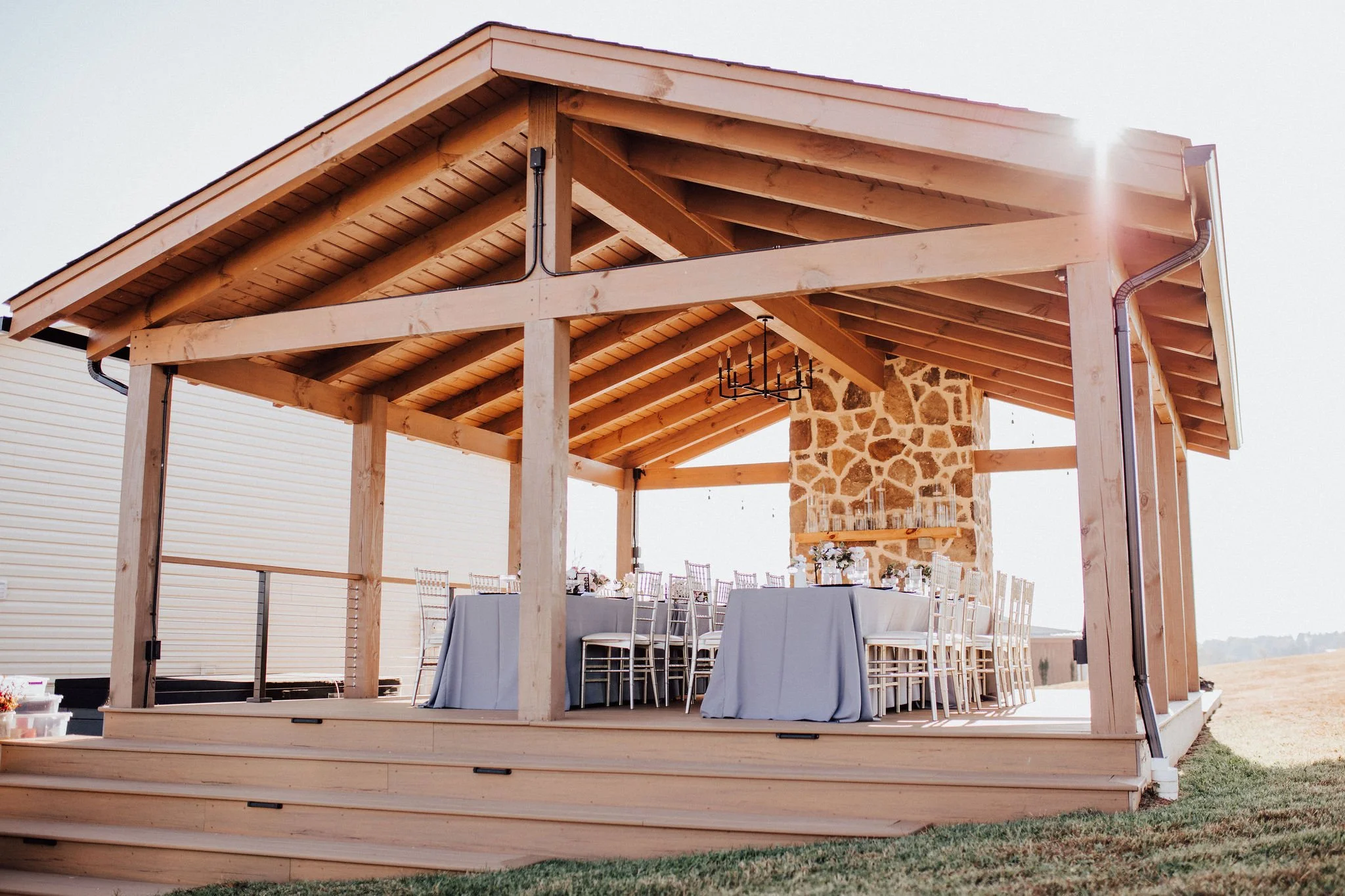 An outdoor pavilion with a wooden frame and a stone fireplace, set up for a dining event with a long table covered with a grey tablecloth, surrounded by white chairs, overlooking a grassy area.
