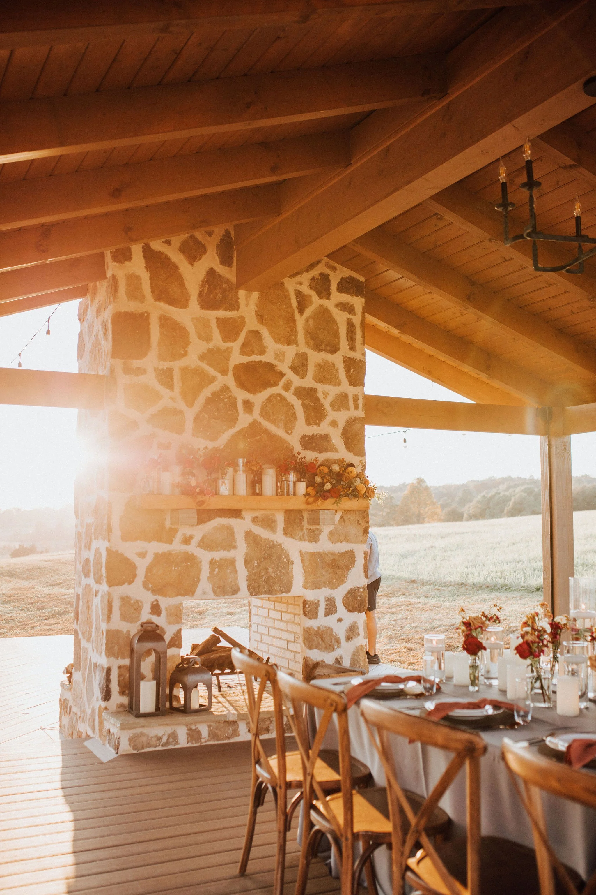 An indoor/outdoor dining area with a stone fireplace, wooden ceiling and beams, decorated for a gathering with floral centerpieces, candles, and place settings on a table. The scene is sunlit, suggesting late afternoon or early evening.