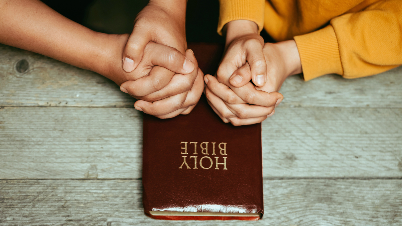 Two people with hands in prayer position resting on the Bible