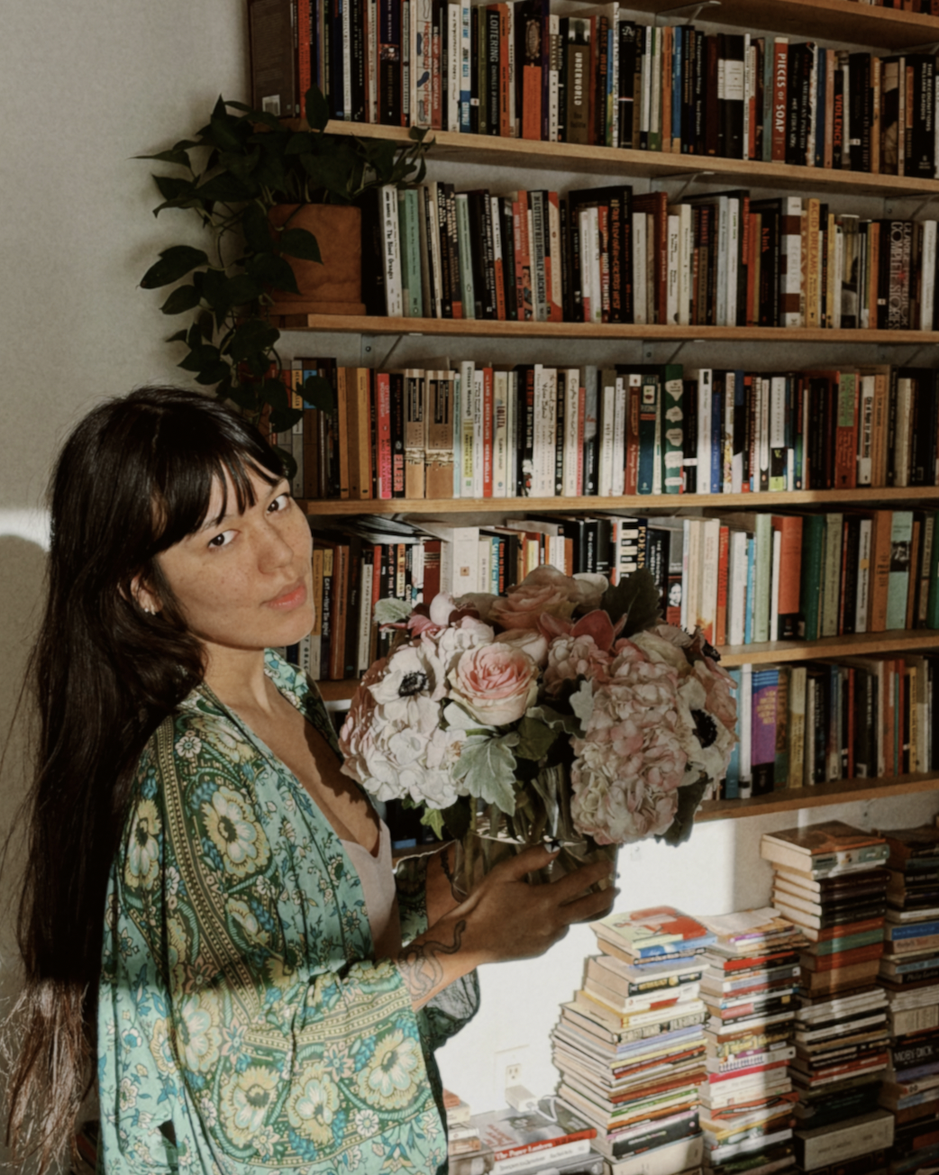 A woman with long dark hair holding a bouquet of pink and white flowers in front of a bookshelf filled with books. She is wearing a green patterned shawl and has a tattoo on her forearm.