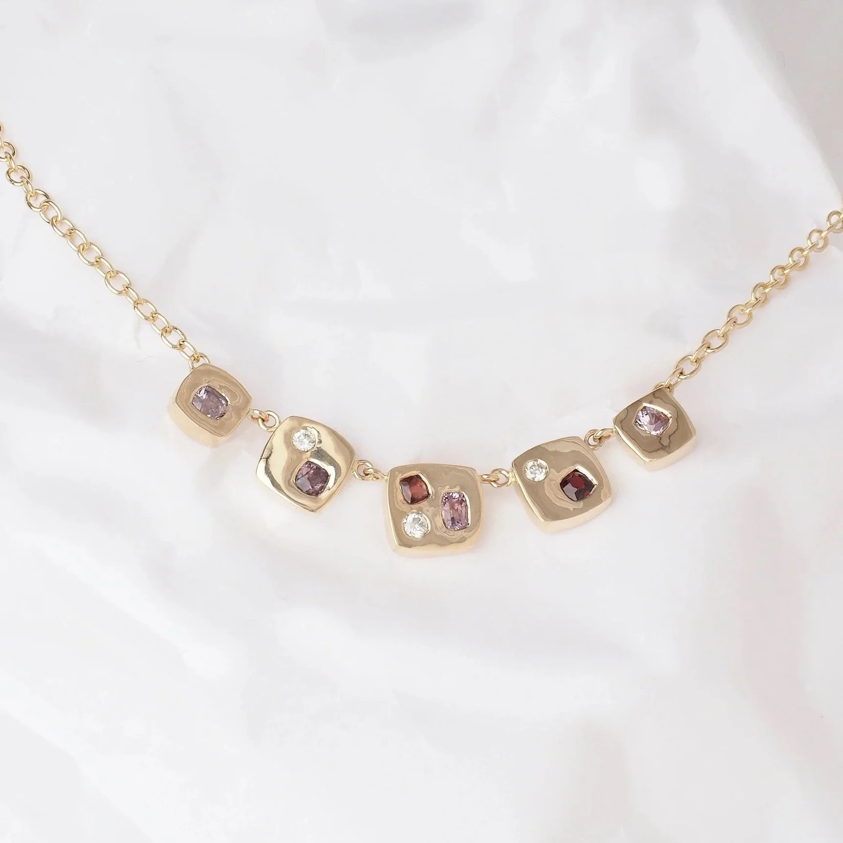 Gold necklace with square and rectangular settings, each containing purple, red, and clear gemstones, on white crinkled paper background.