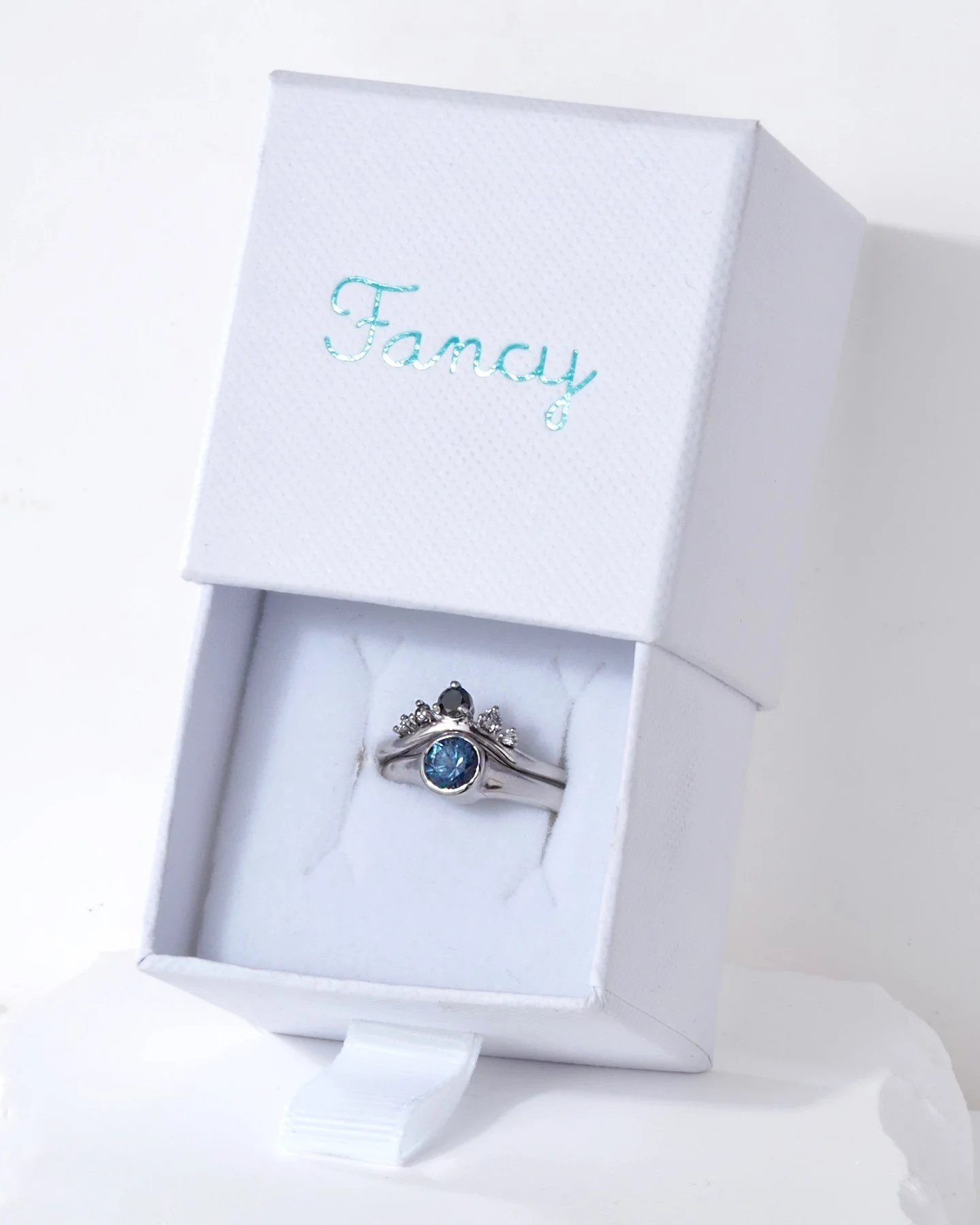 An astonishingly blue sapphire in a fabulously modern bezel setting. The upper ring crowns the sapphire with black and white diamonds on a curved band that brings in the richness of an antique heirloom.  Both rings are made in glossy platinum (recycl
