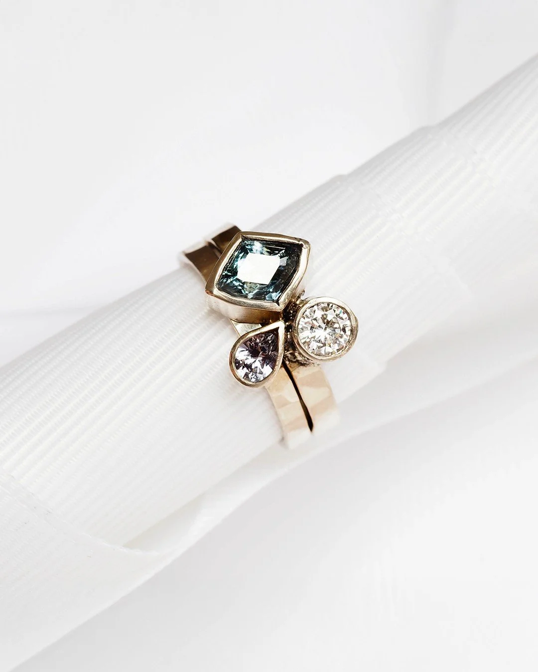 Graceful stacking ring set of two rings, one with a pear-shaped pink tourmaline and the other with a white diamond and a diamond-shaped blue sapphire.

The ring bands are made with recycled 14K gold. The bezel settings are just slightly-organically s