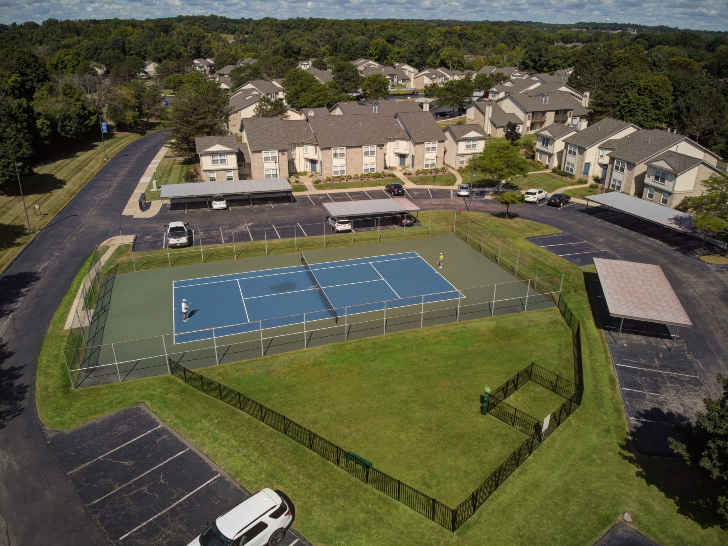 Aerial view of a neighborhood with a tennis court, parking lots, and residential buildings surrounded by trees.
