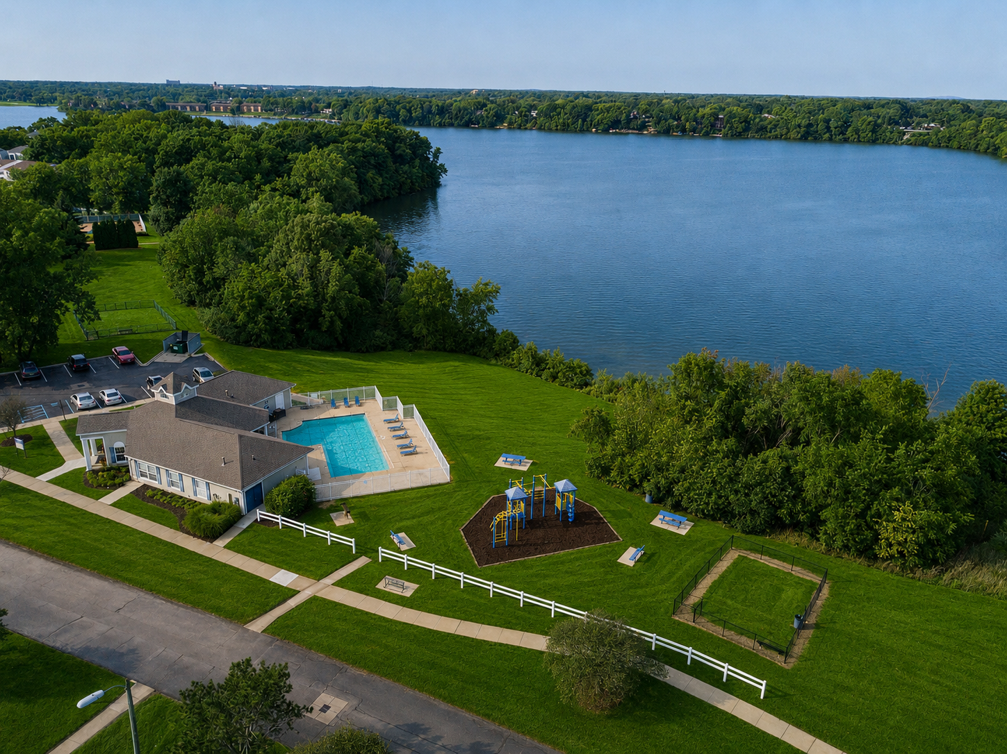 Aerial view of a community pool, playground, and tennis court on green lawns beside a large lake, with parking lot and residential area in the distance.