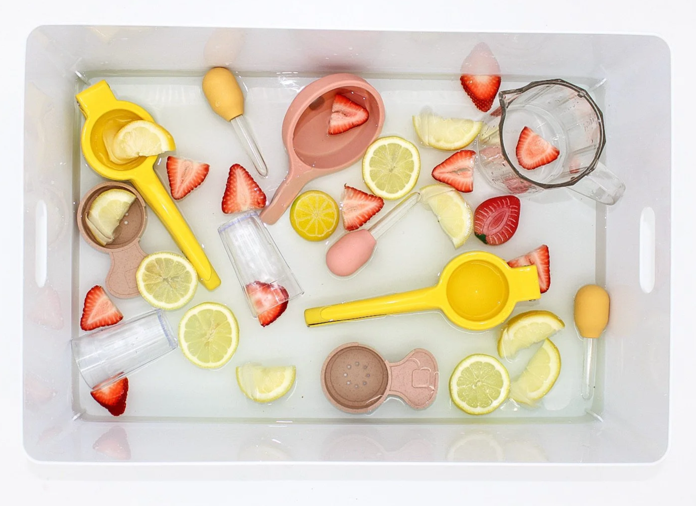 A white tray filled with plastic lemon and strawberry slicers, daiquiri molds, small measuring cups, and lemon and strawberry slices, some of which are floating in water.