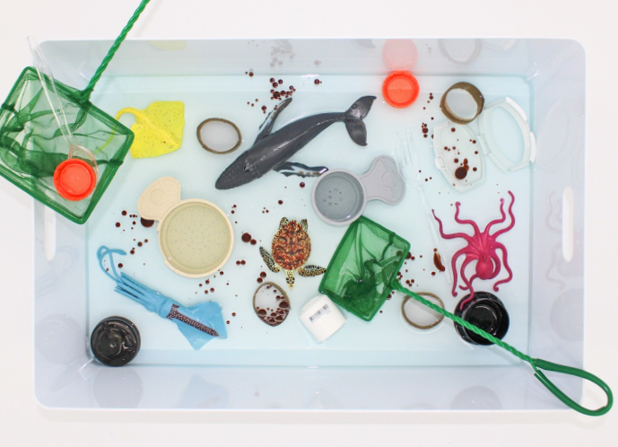 A plastic container filled with water containing small toy sea animals such as a black fish, a purple octopus, and a turtle. There are various plastic objects like a green fishing net, a yellow sponge, and a red and white lifebuoy. Several water ring