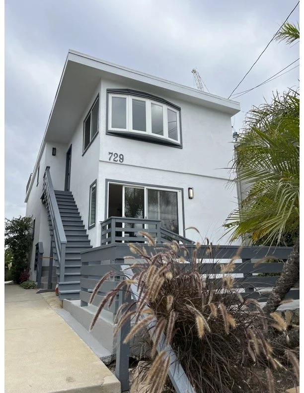 A two-story house with a gray garage door, teal siding, and a gray tiled roof. The house has a balcony on the second floor with a railing, two black-tinted windows, and a double door. There are paint cans and a worker on the sidewalk in front of the house.