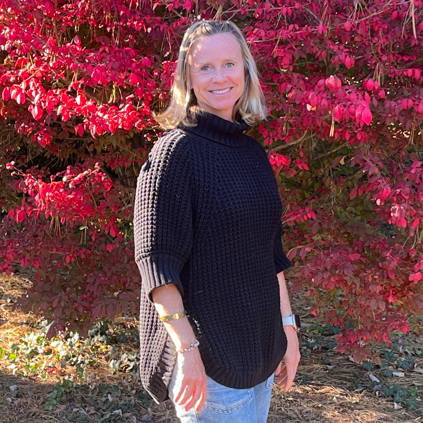 A woman with blonde hair wearing a black sweater standing in front of pink and red foliage.