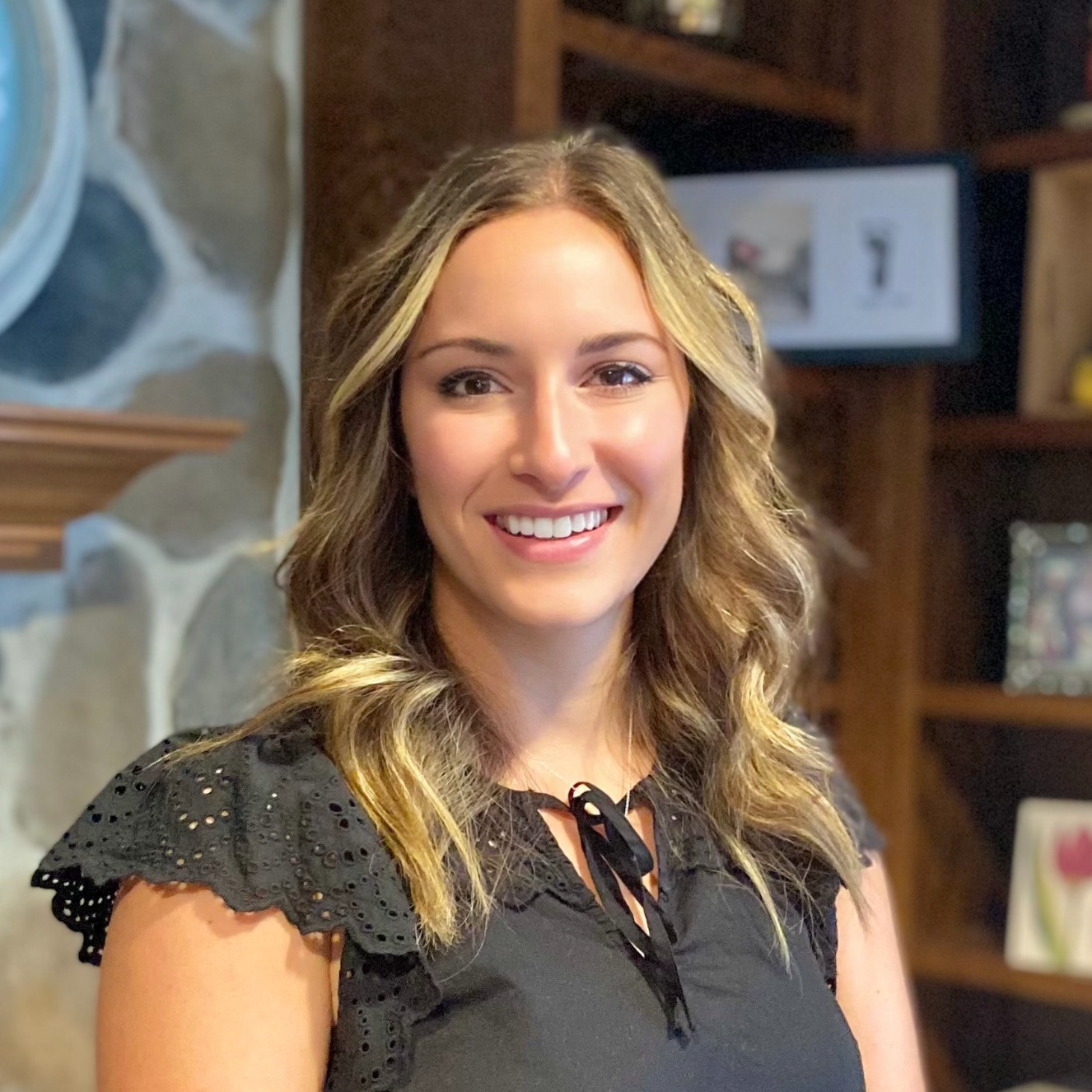 A woman with wavy, blonde hair smiling in front of a stone wall and wooden shelves on a background that includes framed pictures and decorative items.