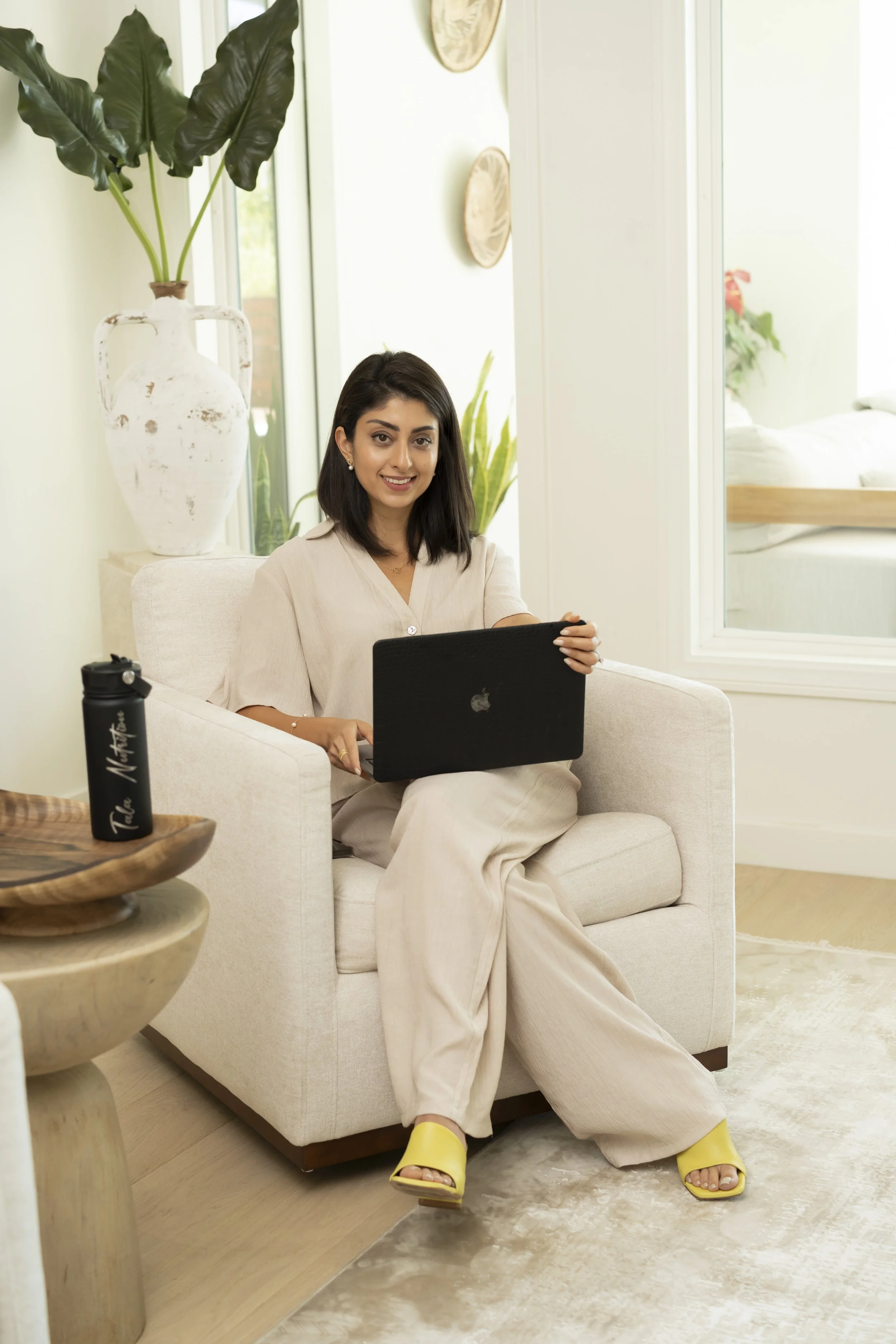 A woman sitting on a white armchair working on a black laptop in a bright, modern living room with large windows, decorative plants, and light-colored decor.