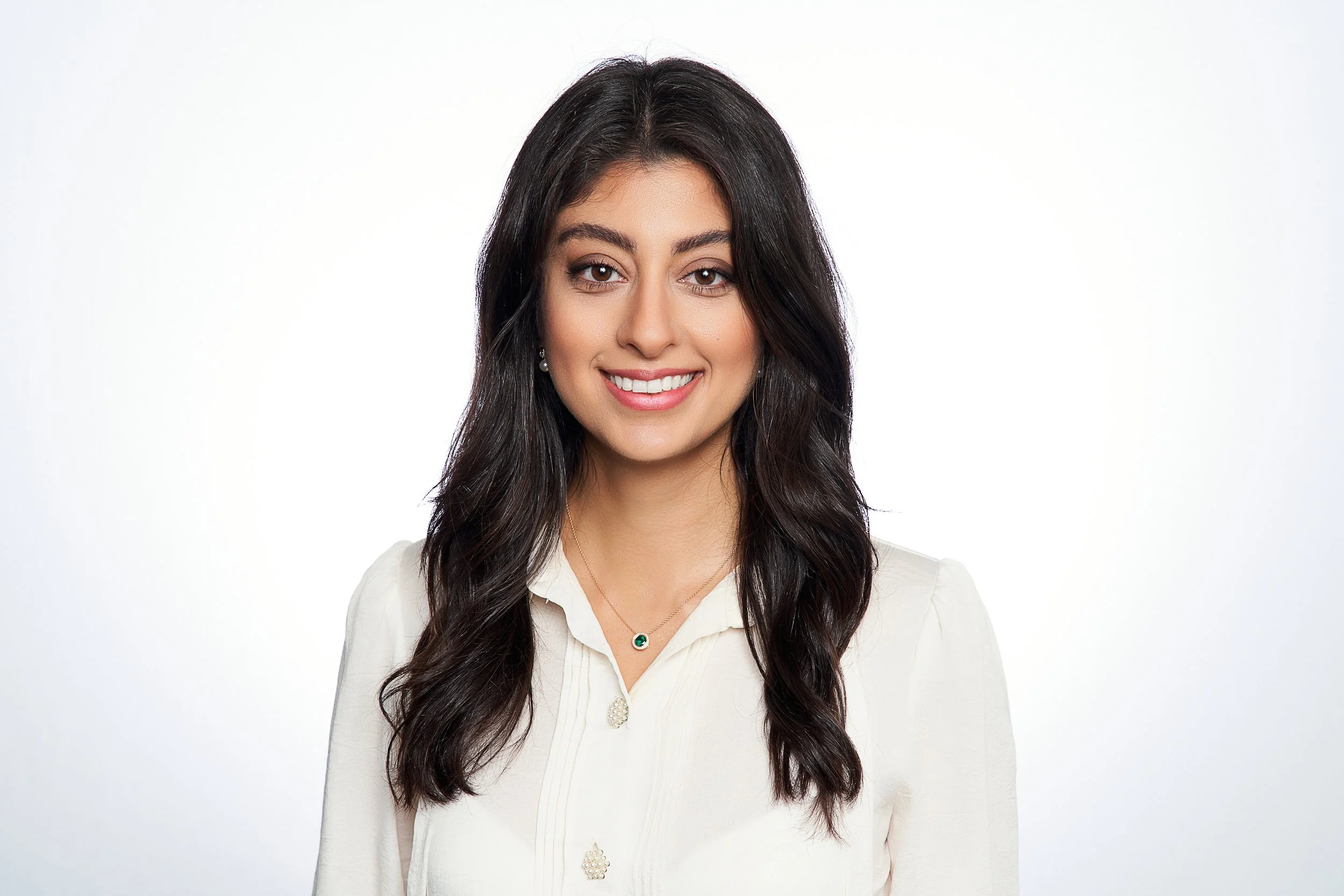 A young woman with long dark hair smiling, wearing a cream-colored blouse and a necklace with a green pendant, against a white background.