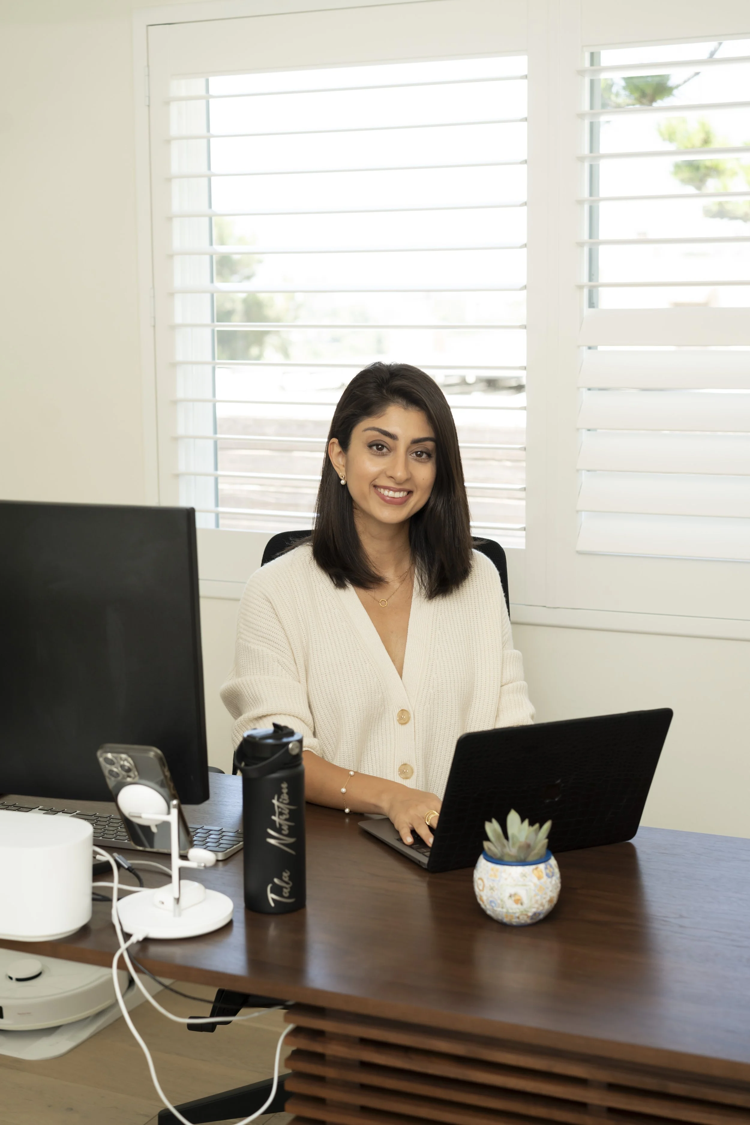 A woman with shoulder-length dark hair sitting at a wooden desk with a laptop, desktop monitor, and a succulent plant, smiling at the camera in a bright office with large windows.