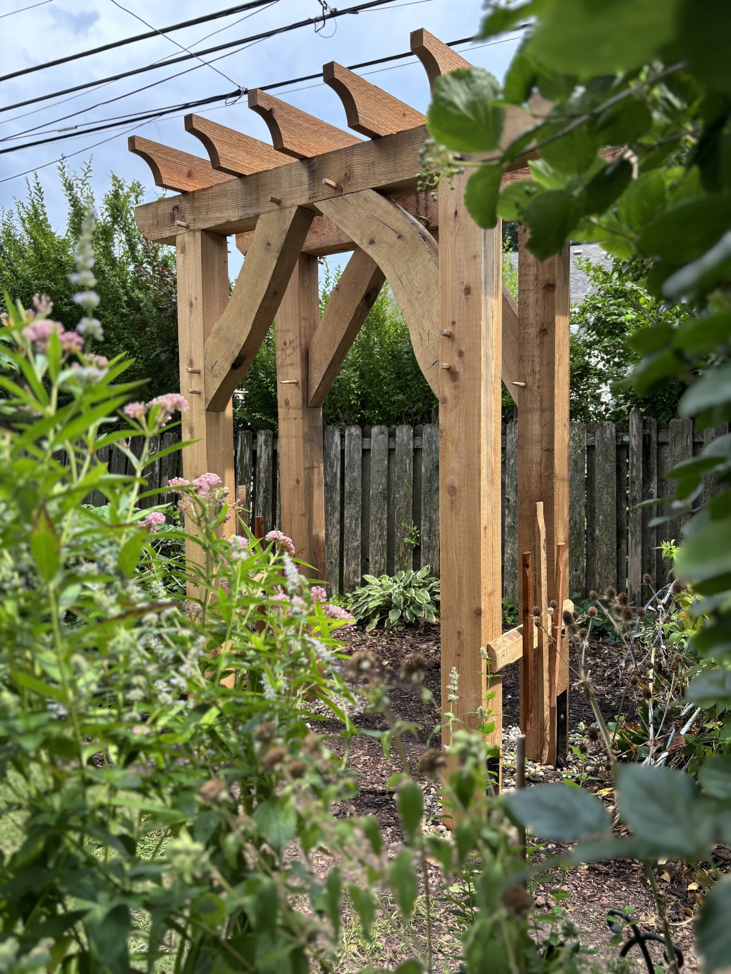 Wooden garden arbor under construction in a garden with flowering plants and a wooden fence in the background.