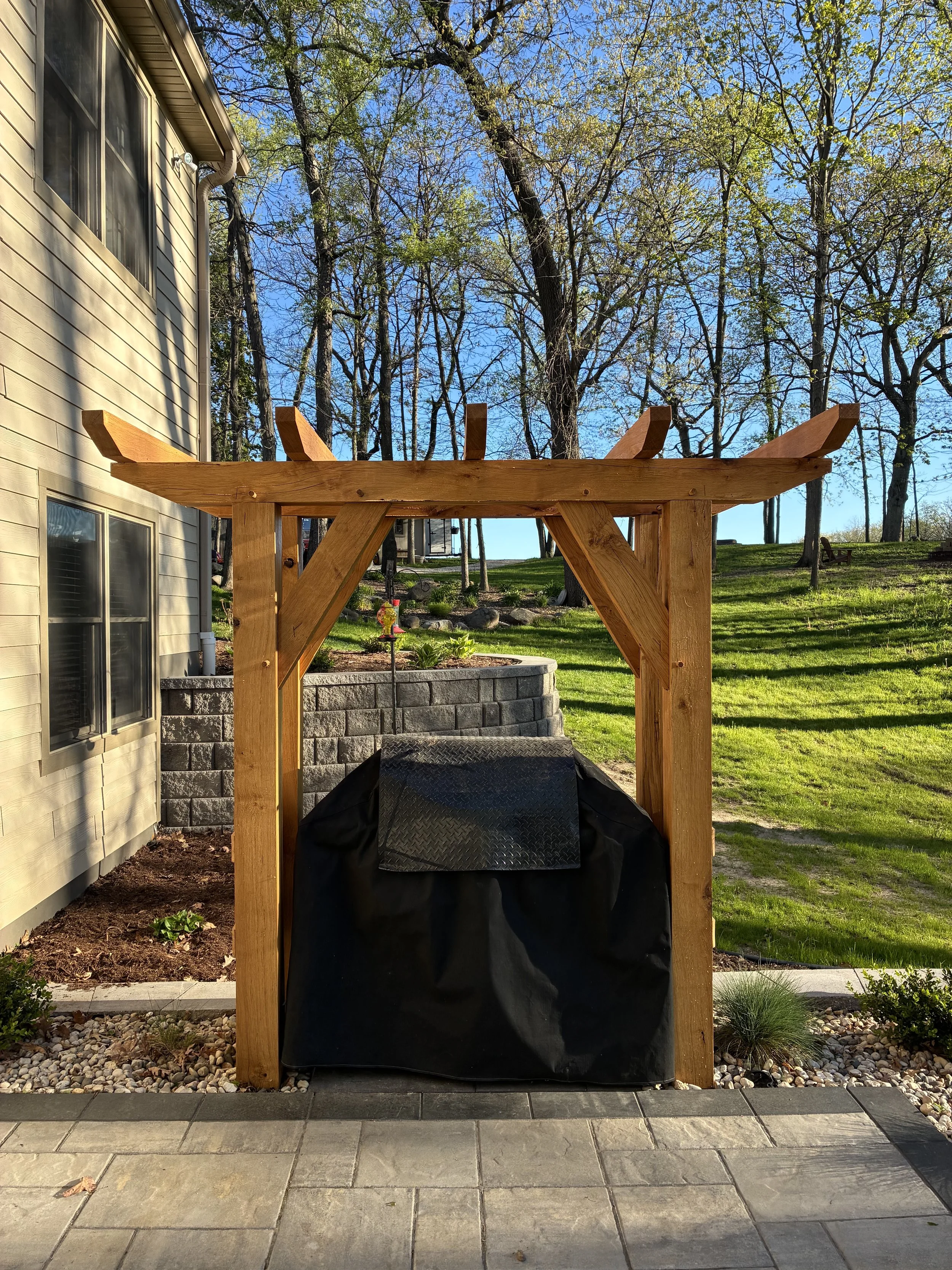 A wooden pergola structure with a black covered barbecue grill underneath, located in a backyard with green grass, trees, and a brick retaining wall. The house siding is visible on the left, and the sky is blue with some clouds.