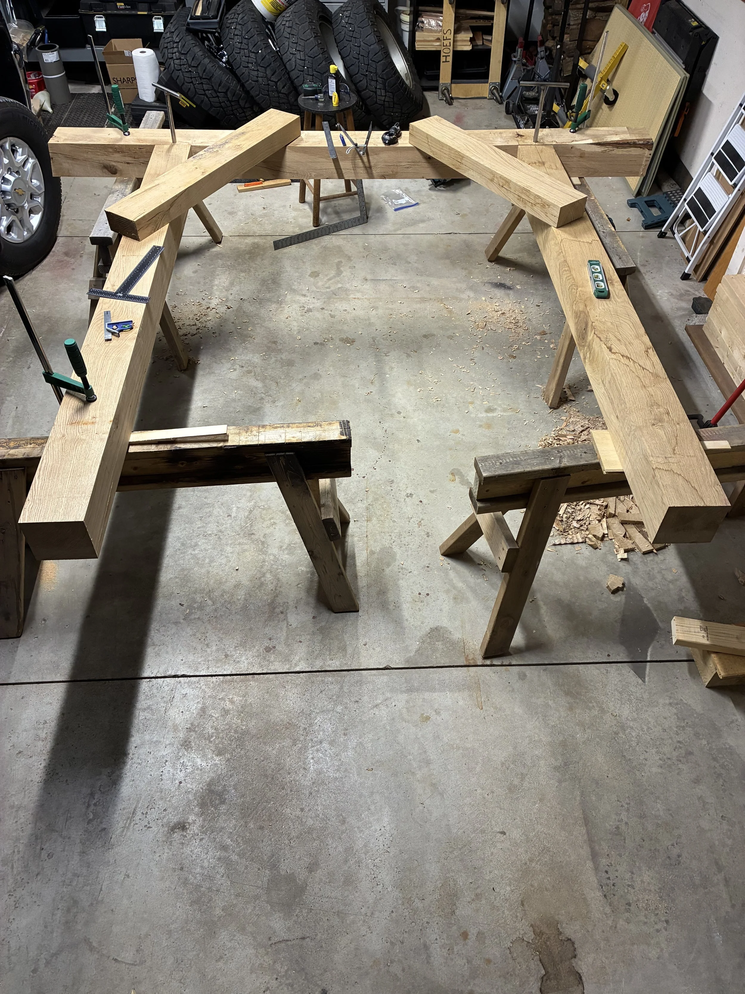 A woodworking project in progress showing a large, roughly rectangular wooden frame assembled and clamped on sawhorses in a garage workshop. Various tools and equipment are scattered around, including clamps, a level, and a measuring tape.