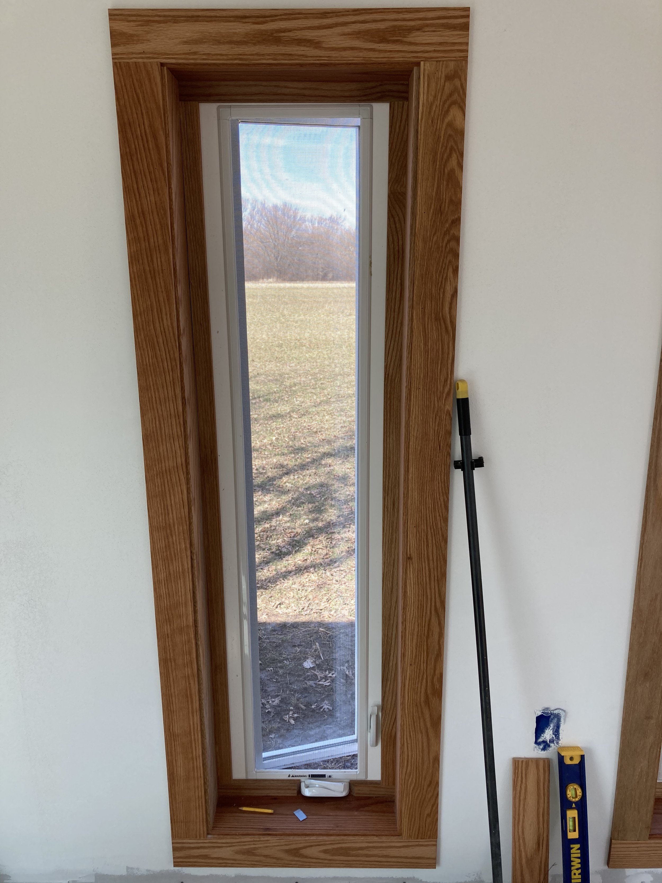 A window with a wooden frame, showing an outdoor landscape with grass and trees, and tools including a level and a measuring stick leaning against the wall.