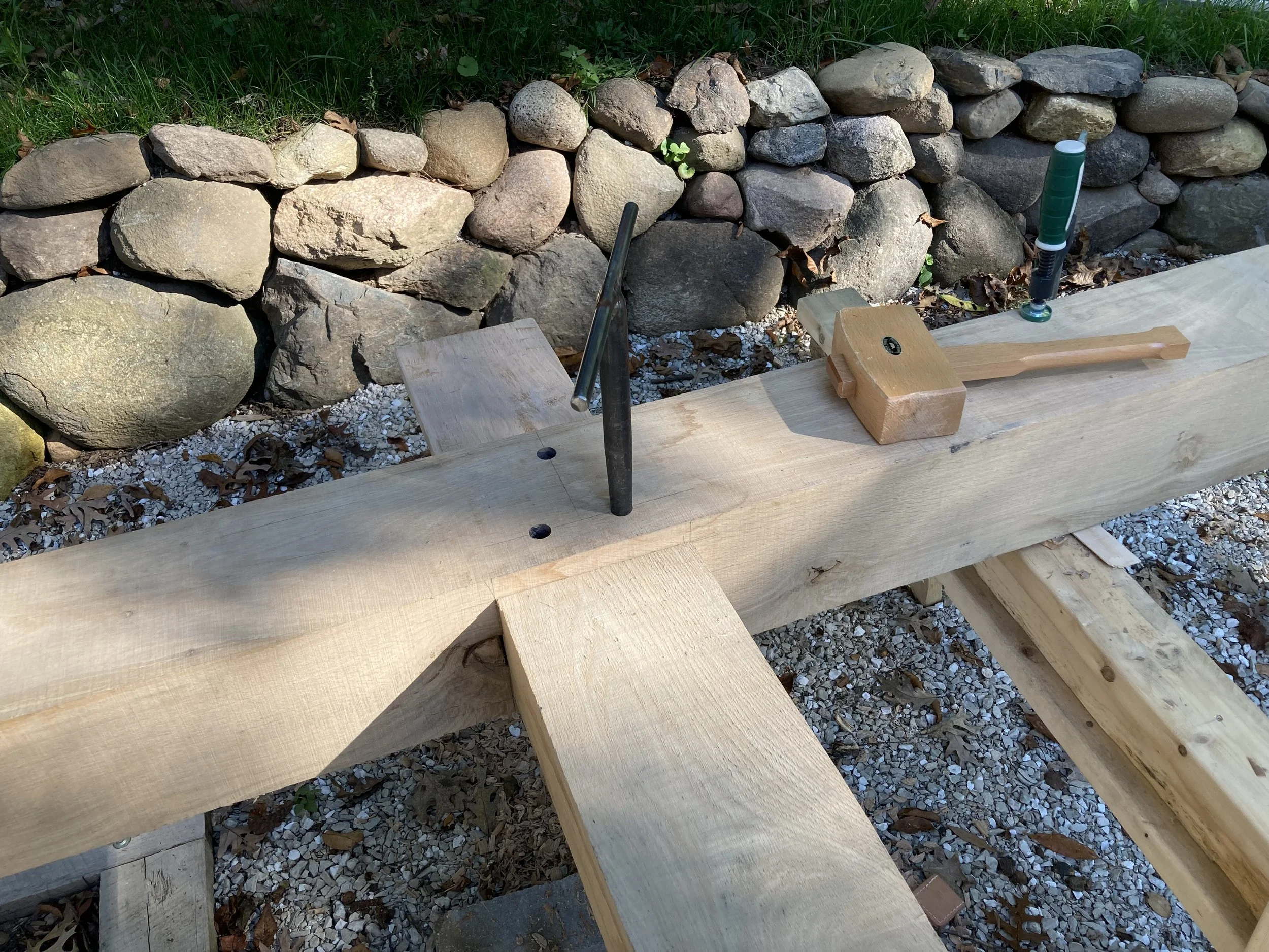 Wooden beam on a workbench with tools including a hammer and screws, next to a stone wall and gravel ground.
