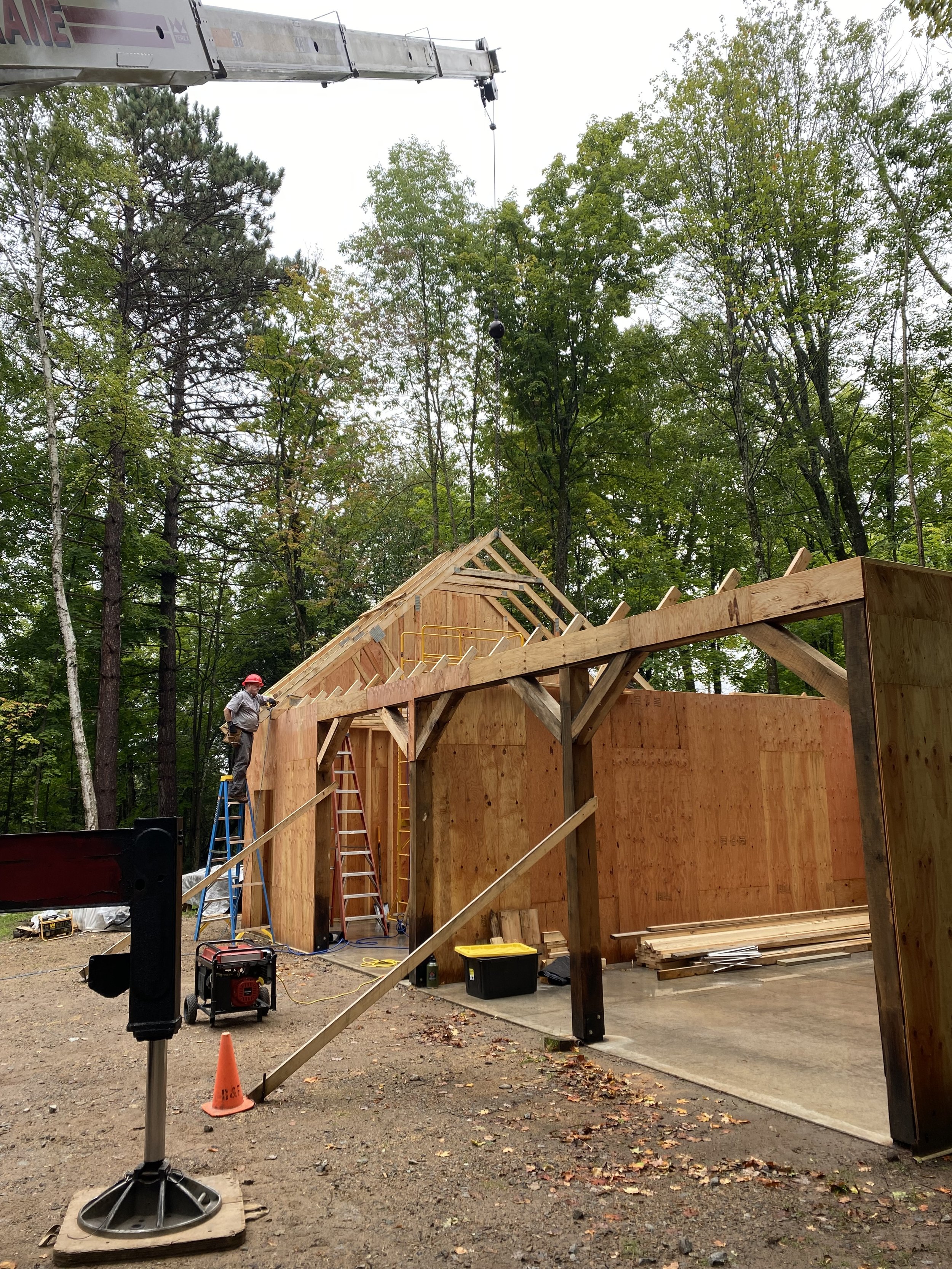 Construction site with a partially built wooden structure surrounded by trees, a worker in a red helmet, various construction tools and equipment, including ladders, a crane in the background, and safety cones.