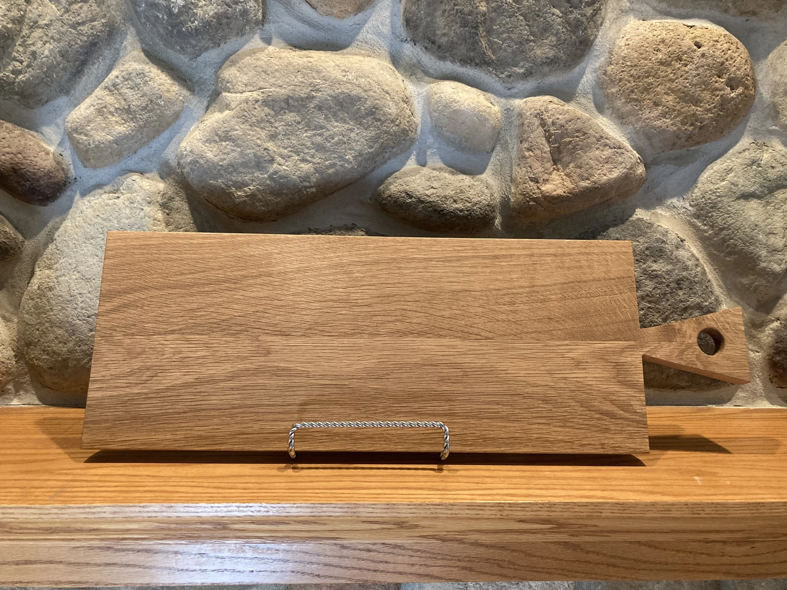 A rectangular wooden cutting board with a circular hole in the handle, resting on a wooden surface against a stone wall background.
