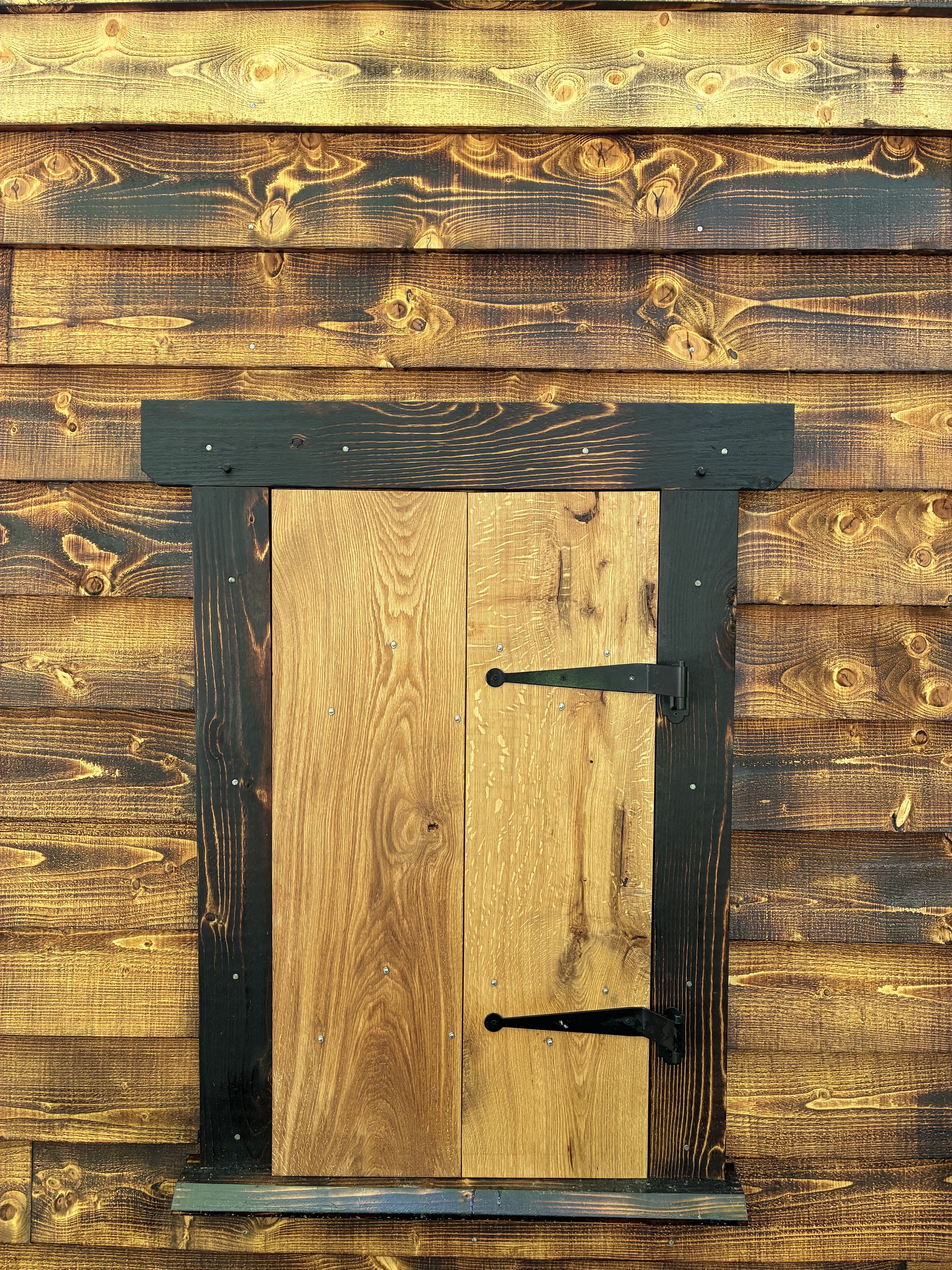 A small wooden door with metal hinges, set into a larger wooden wall, painted in natural and dark stain colors.