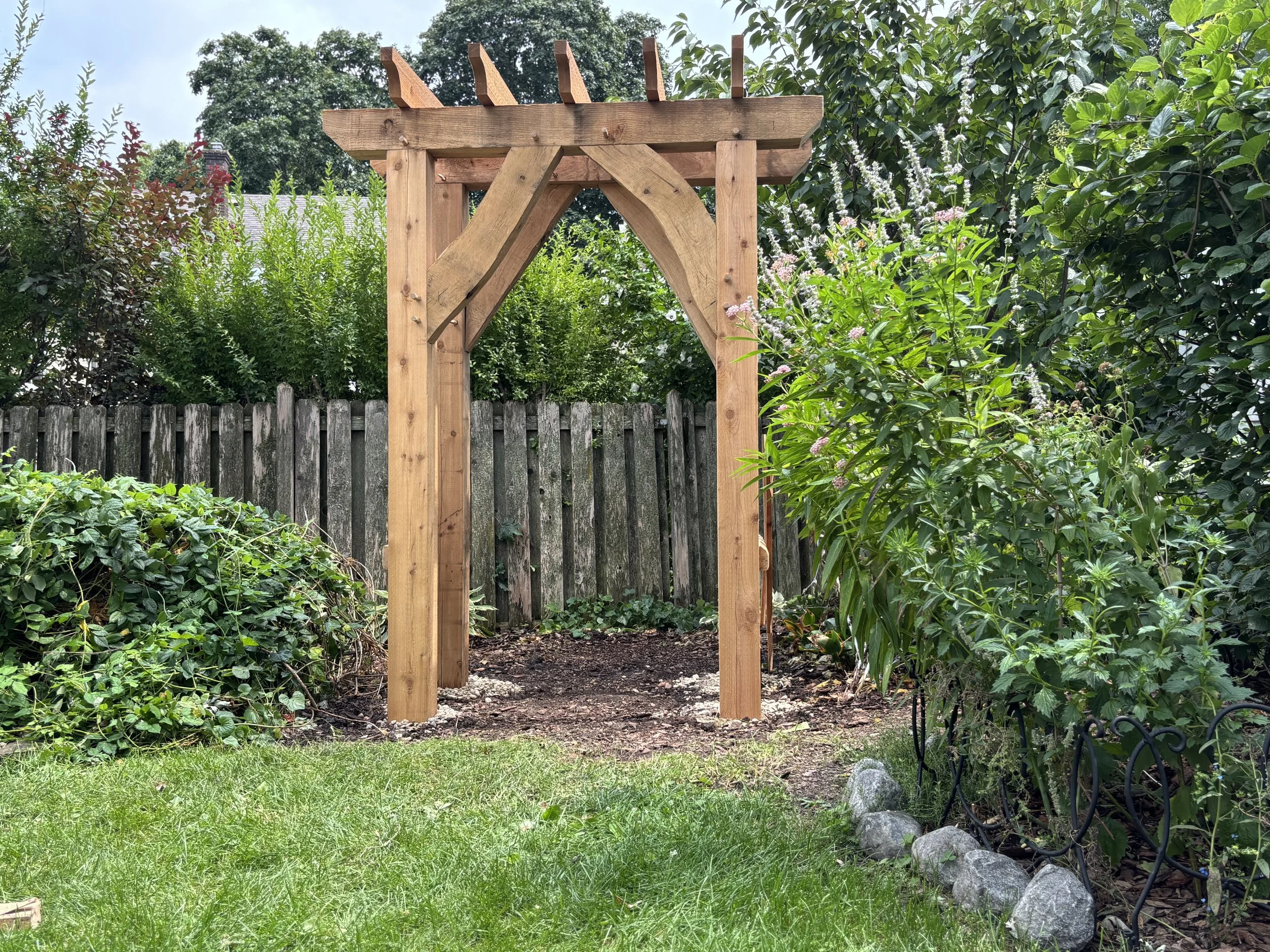 Wooden garden arbor with an arch and slats on top, surrounded by green bushes, a wooden fence, and a grassy lawn.