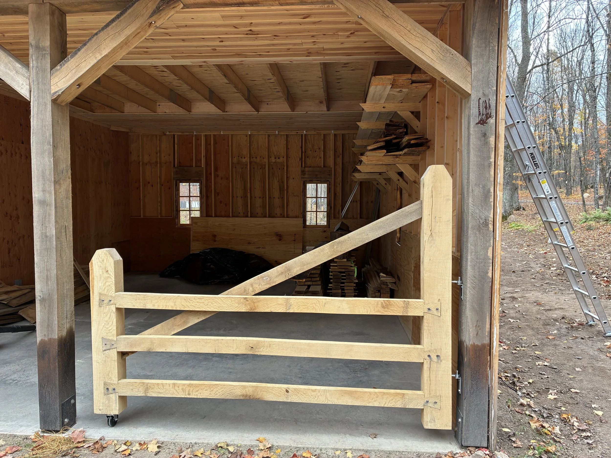 Inside view of a wooden garage under construction with unfinished walls and ceiling, two small windows, a wooden workbench, stacked wood pieces, a black plastic bag, and a ladder outside leaning against the garage entrance.