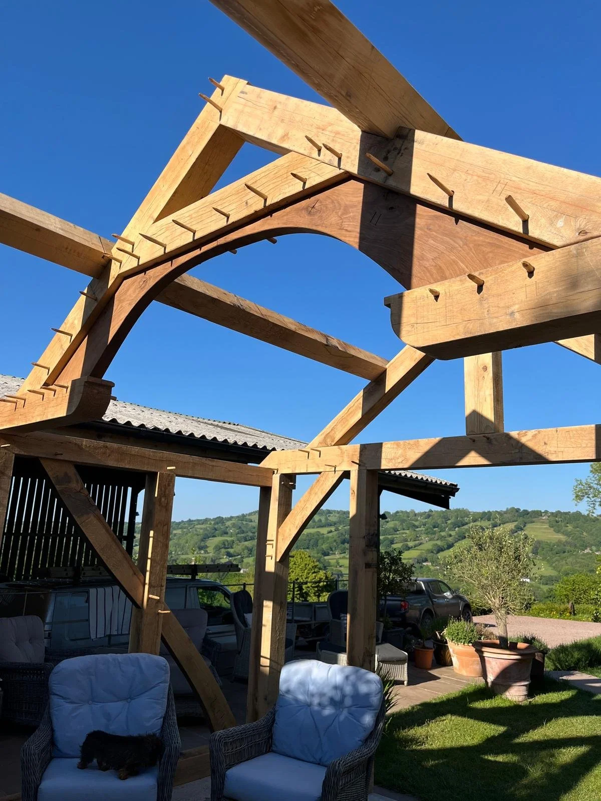 Construction of a wooden outdoor structure with a clear blue sky, green hills, and parked cars in the background. Patio furniture including white cushioned chairs and potted plants are in the foreground.