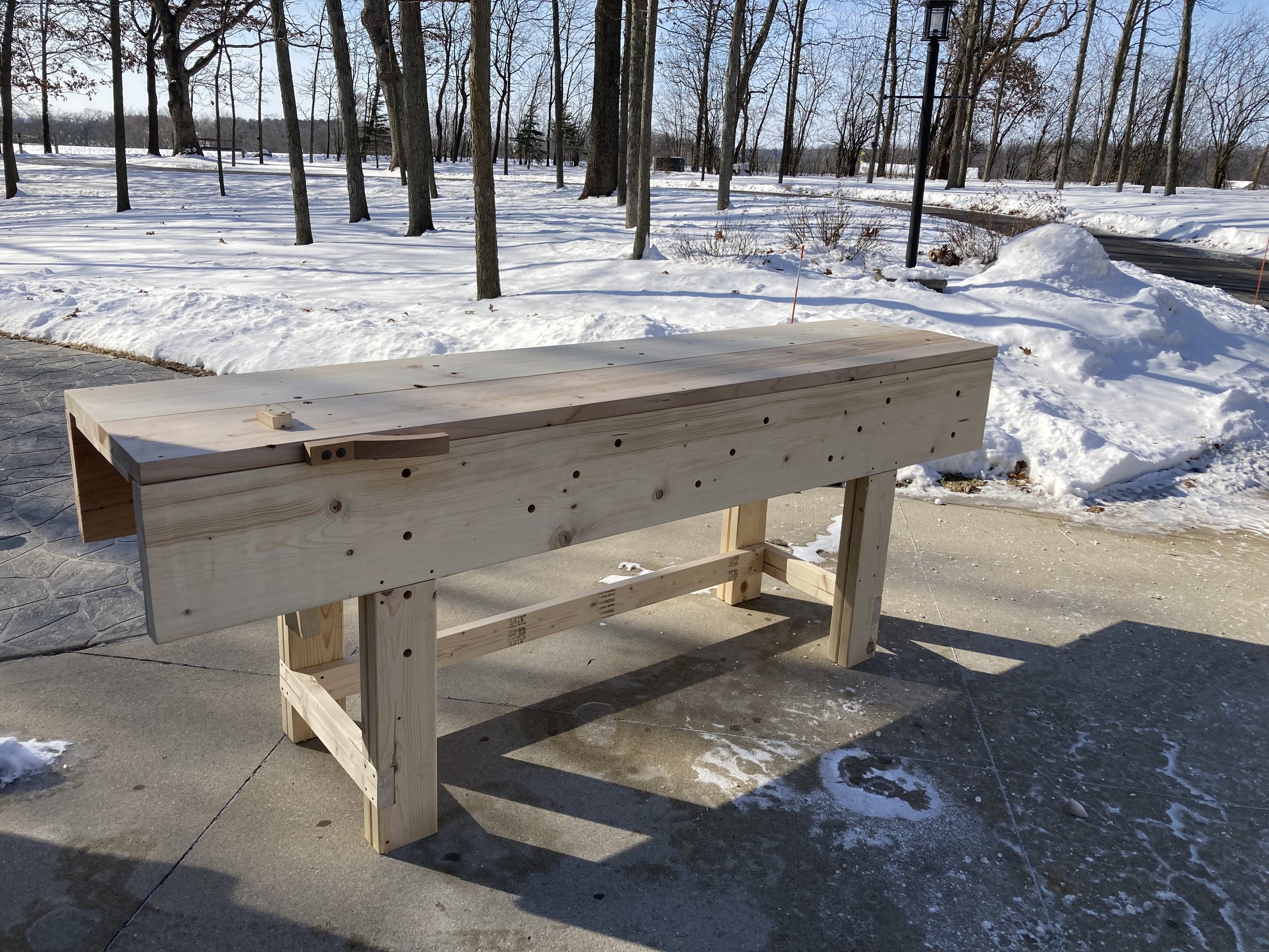 A wooden workbench is outdoors on a concrete patio with snow and leafless trees in the background during winter.