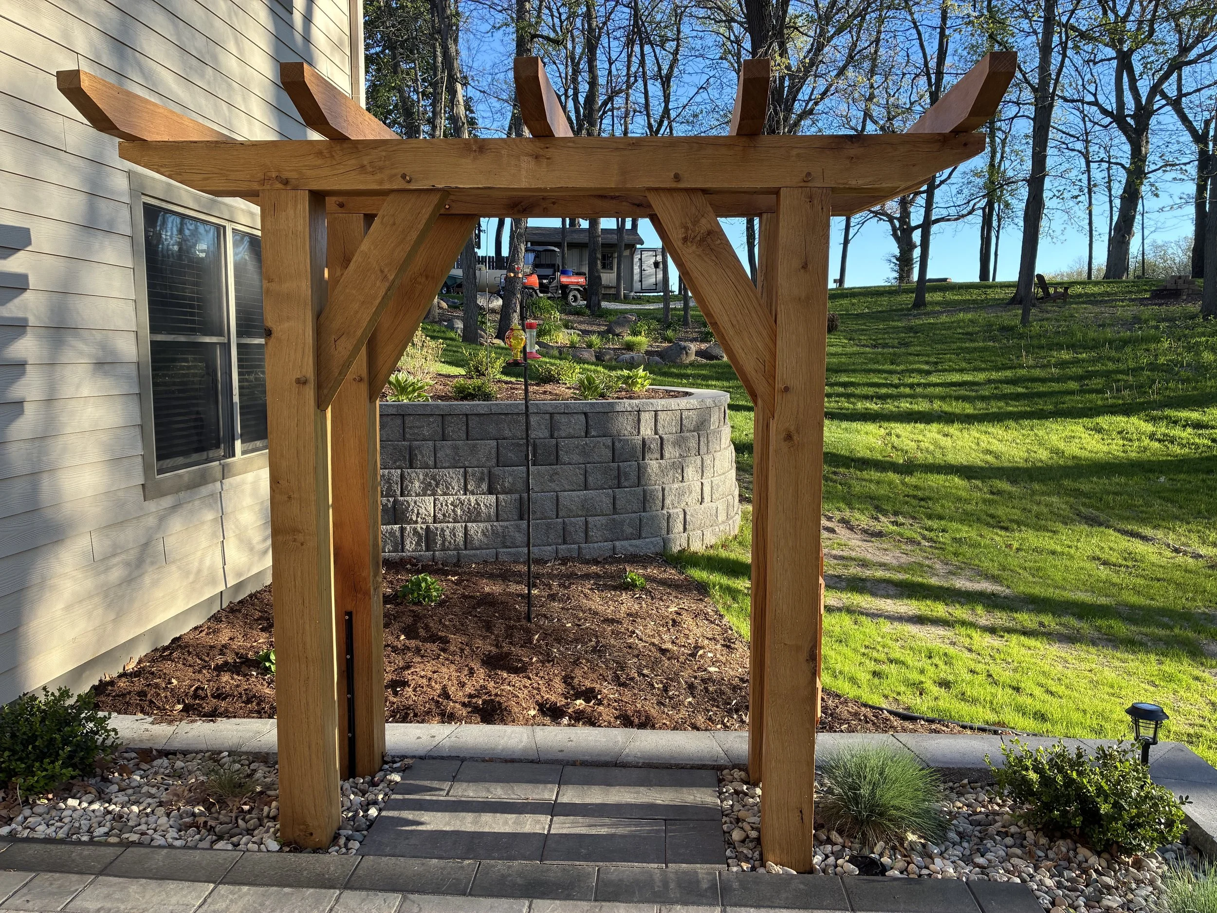 Wooden pergola structure next to house with window, surrounding landscape includes mulched flower bed, small bushes, and trees with a grassy yard in the background.
