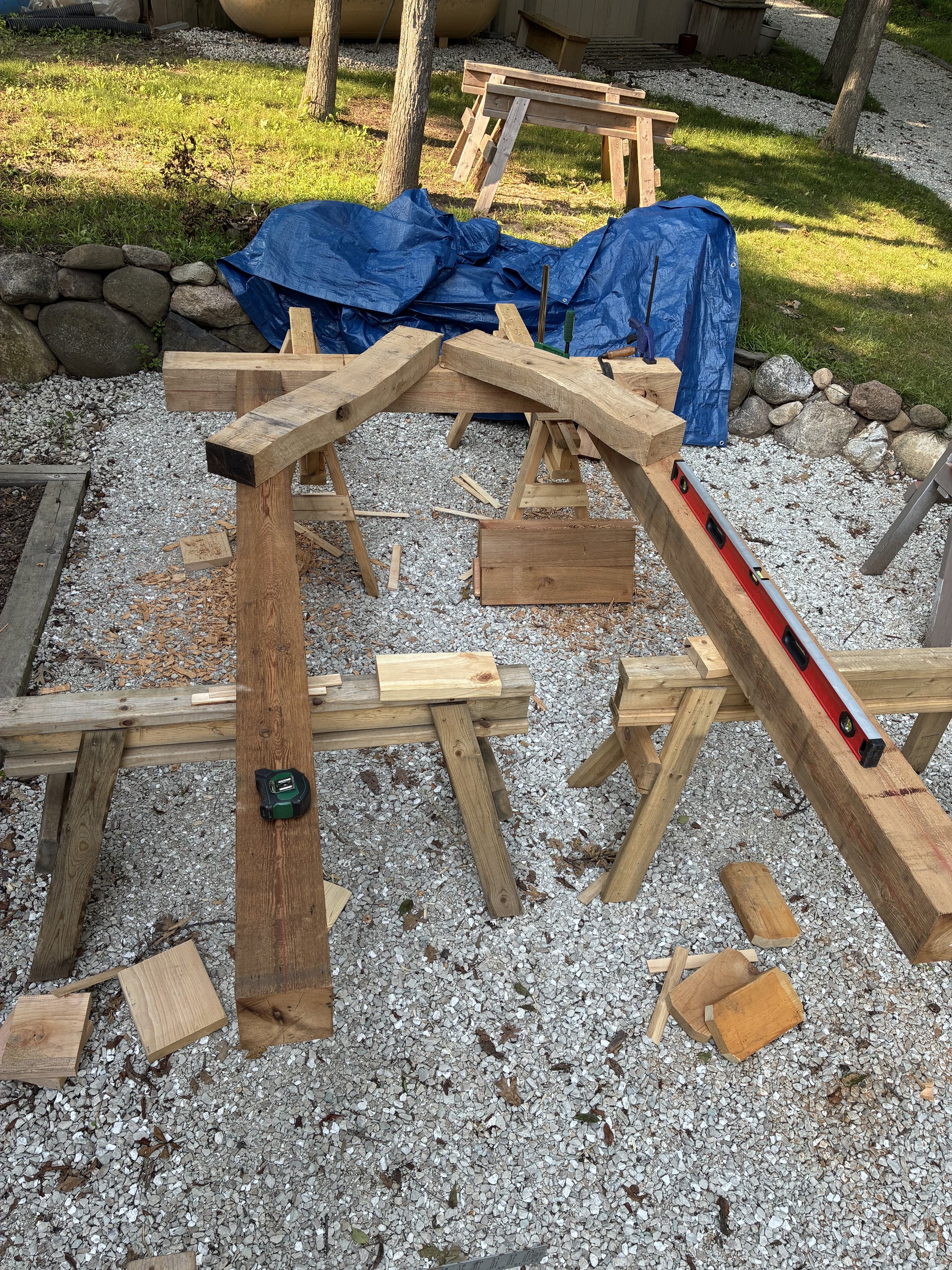A woodworking workspace outdoors with various wood pieces, tools, and sawhorses on a gravel surface, with trees and grass in the background.