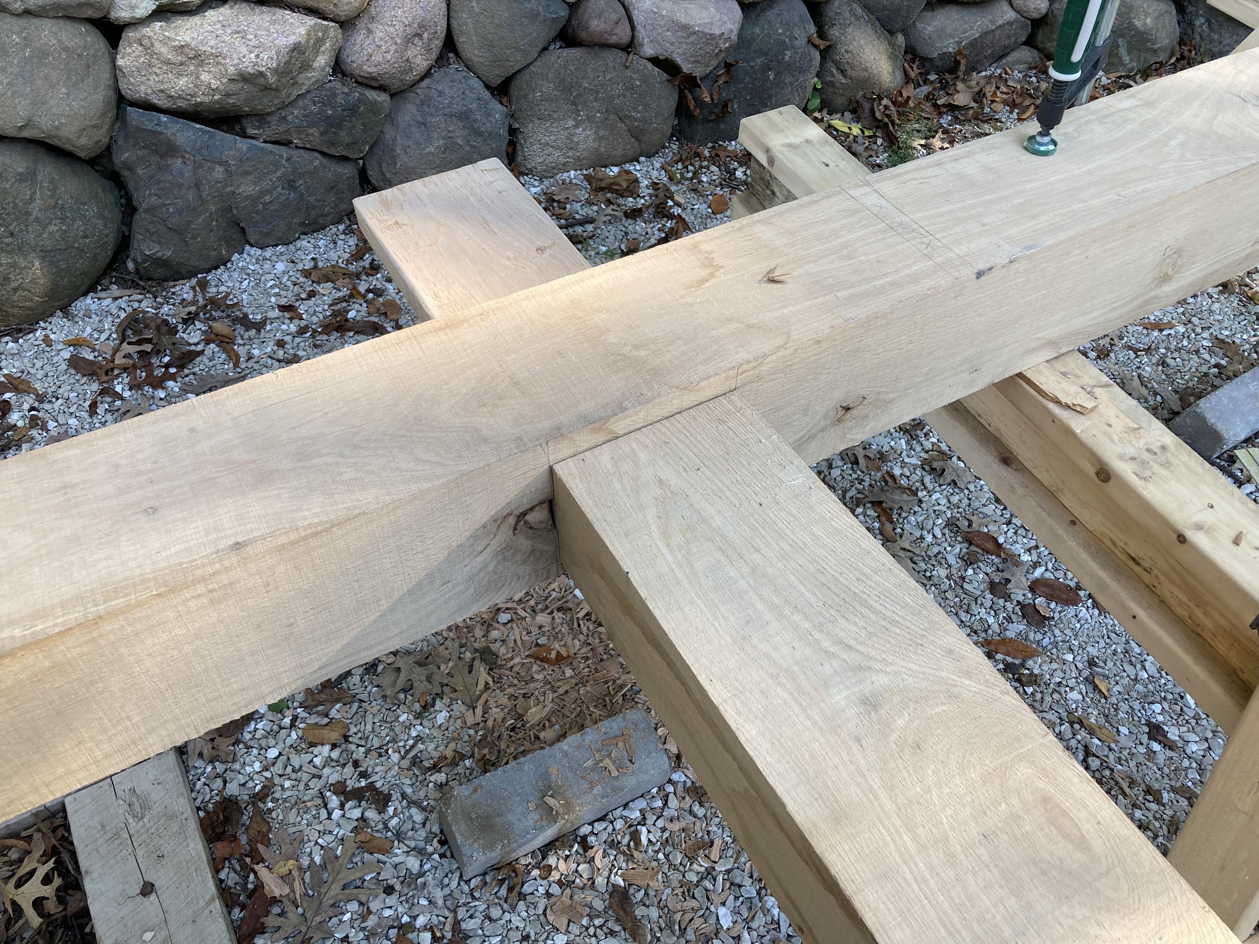 Close-up view of a woodworking project showing several large, cut pieces of wood assembled on a gravel surface with a stone wall in the background. A screw is partially driven into one of the pieces of wood.